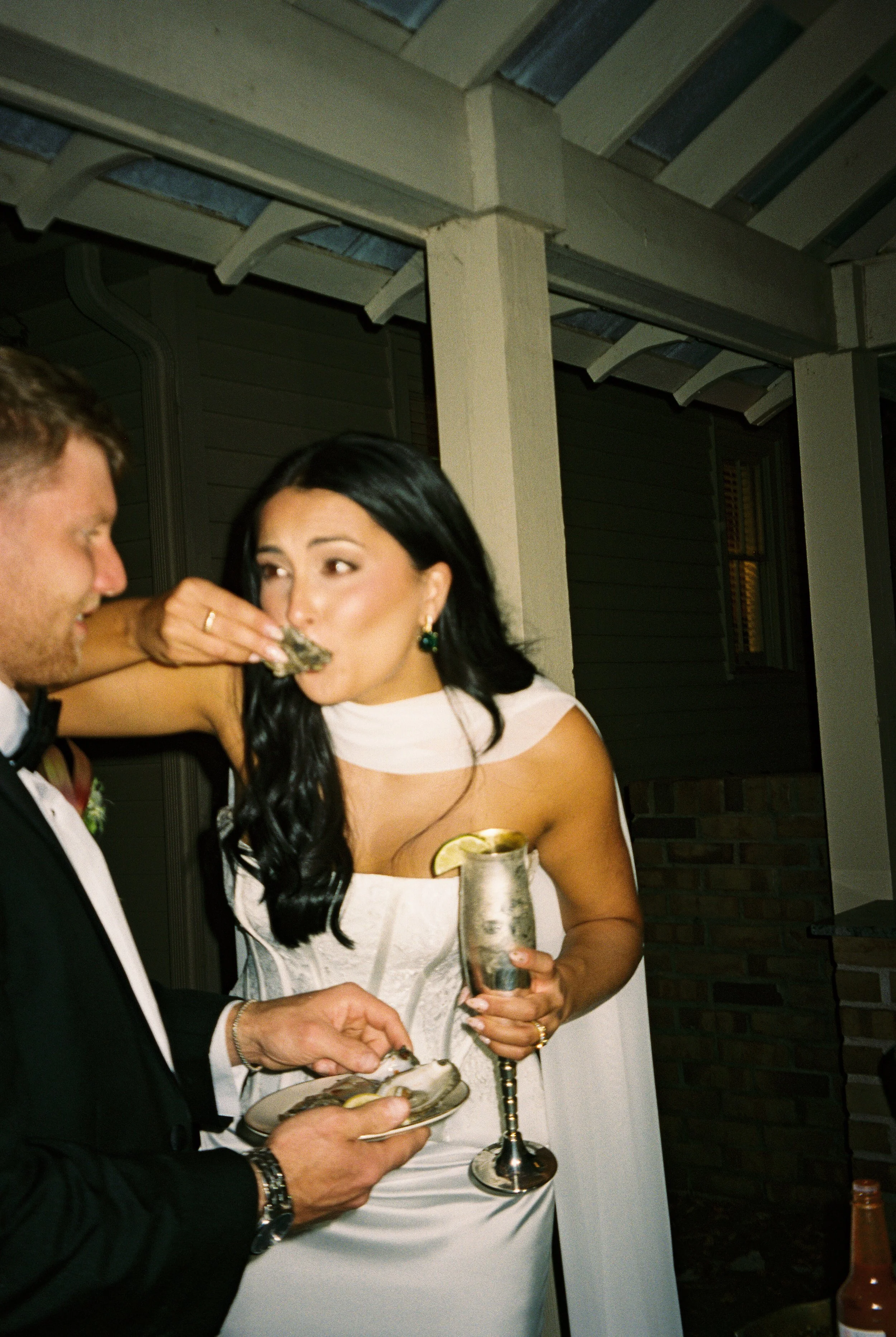 Bride eating an oyster while holding a silver champagne flute during a candid nighttime wedding reception.
