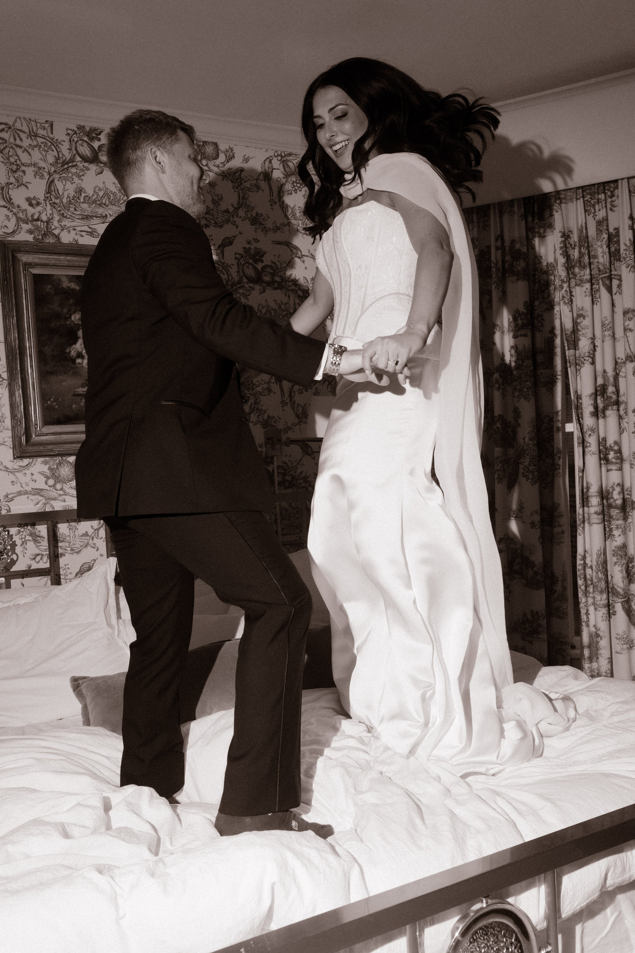 Black-and-white wedding photograph of a bride and groom laughing and dancing together on a bed.