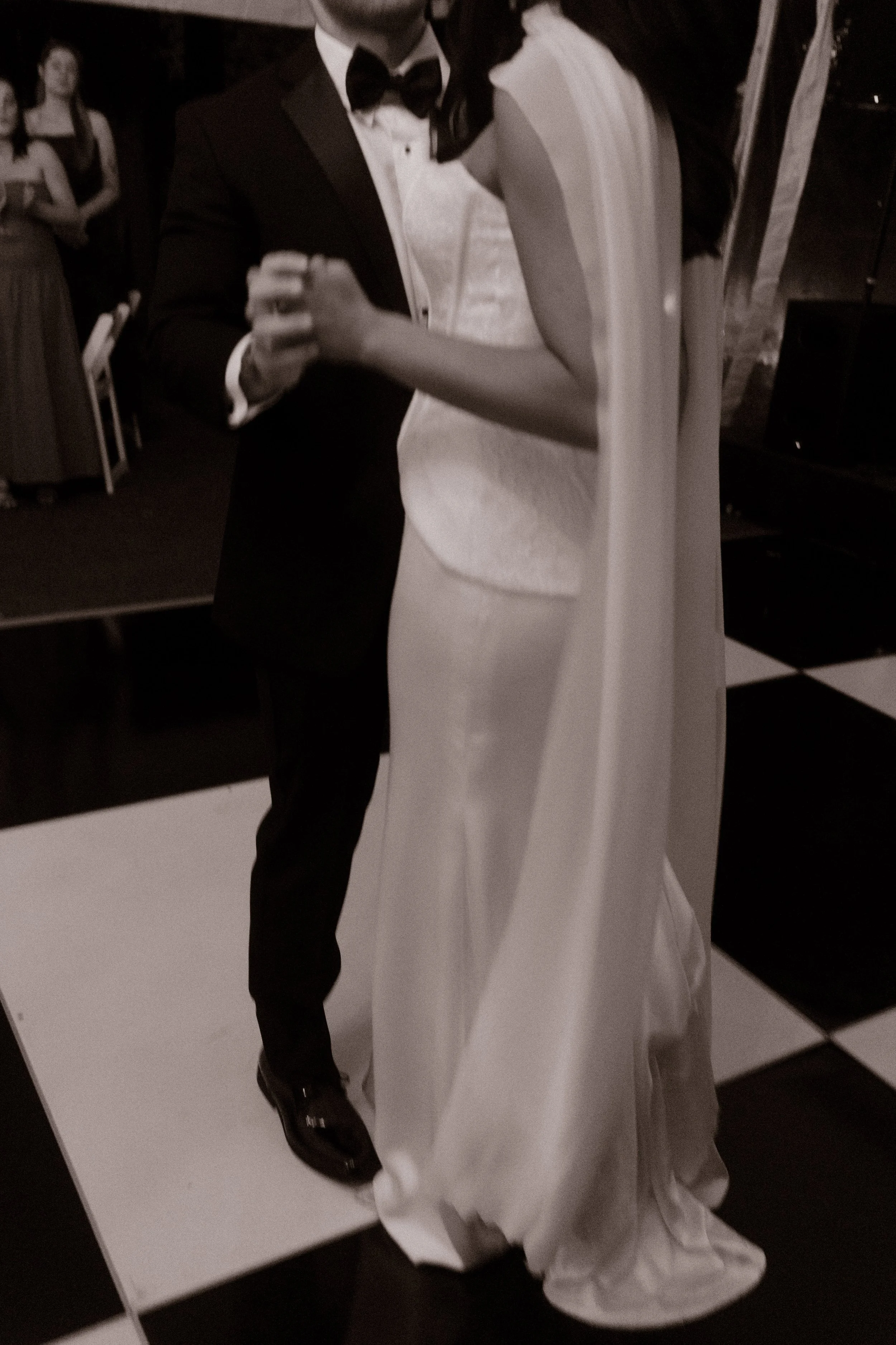 Black-and-white documentary wedding photo of a bride and groom sharing a slow dance on a checkered dance floor.