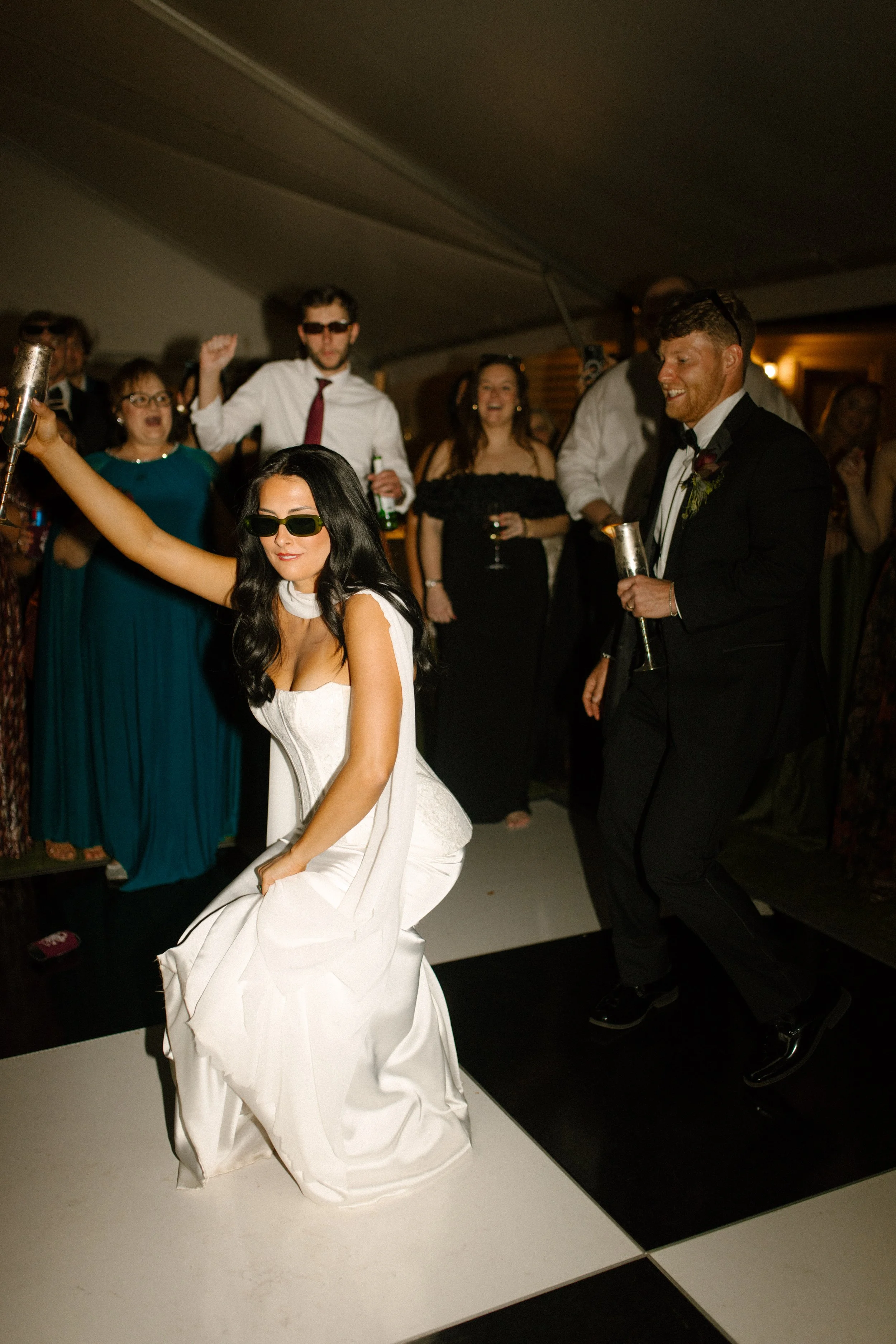 Bride dancing in sunglasses on a black-and-white reception dance floor, surrounded by guests and moody wedding colors.