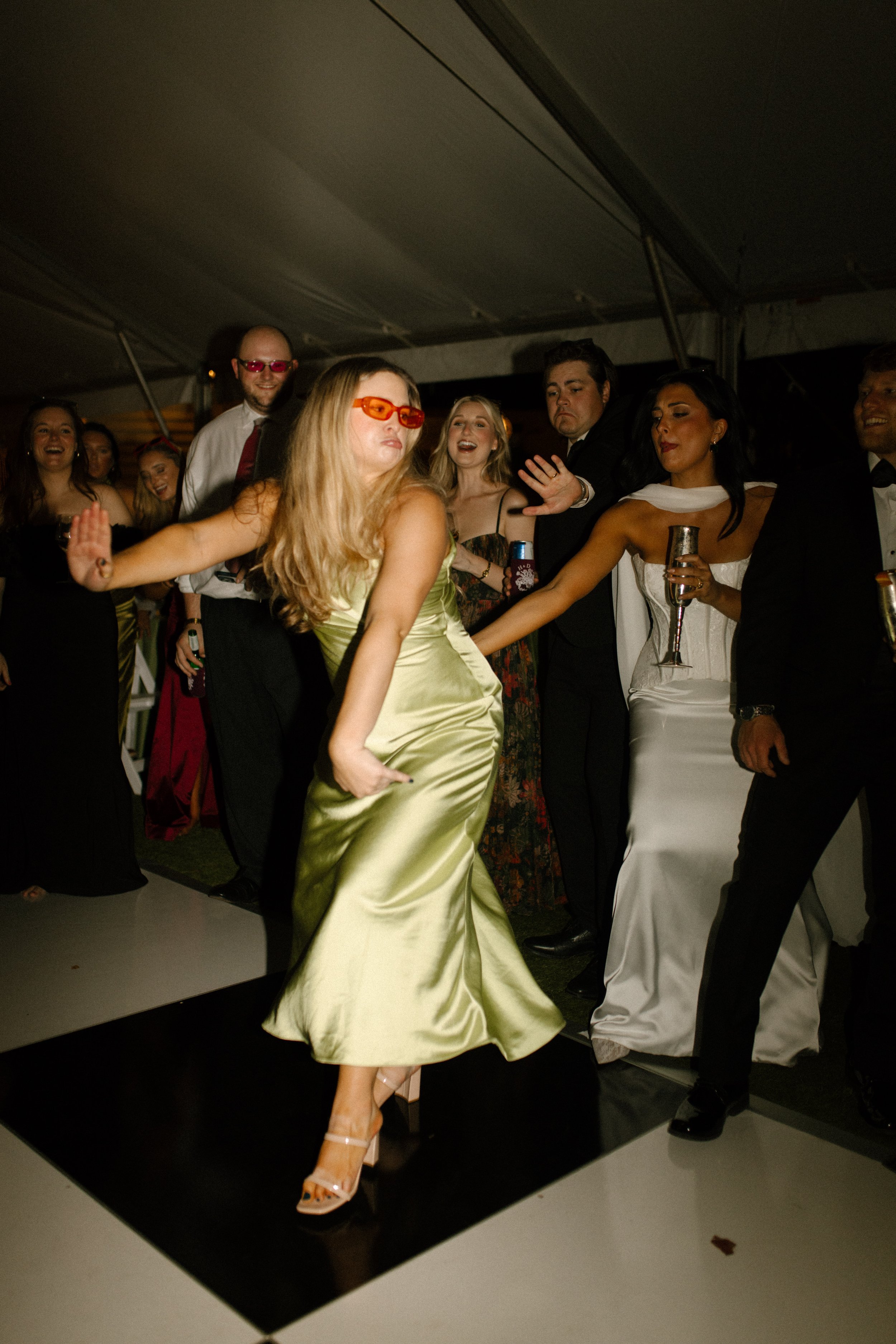 Wedding guest dancing in a light green satin dress and orange sunglasses while the bride celebrates nearby.