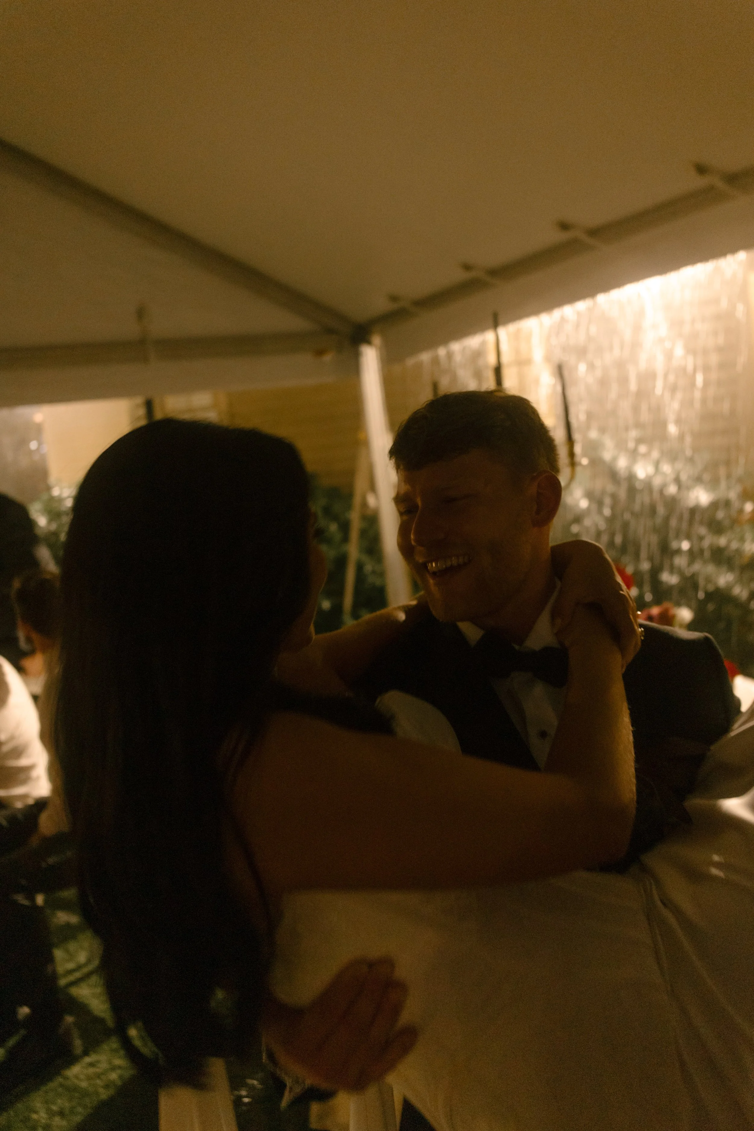 Bride and groom embracing beneath warm reception lighting, with rain falling outside their wedding tent.