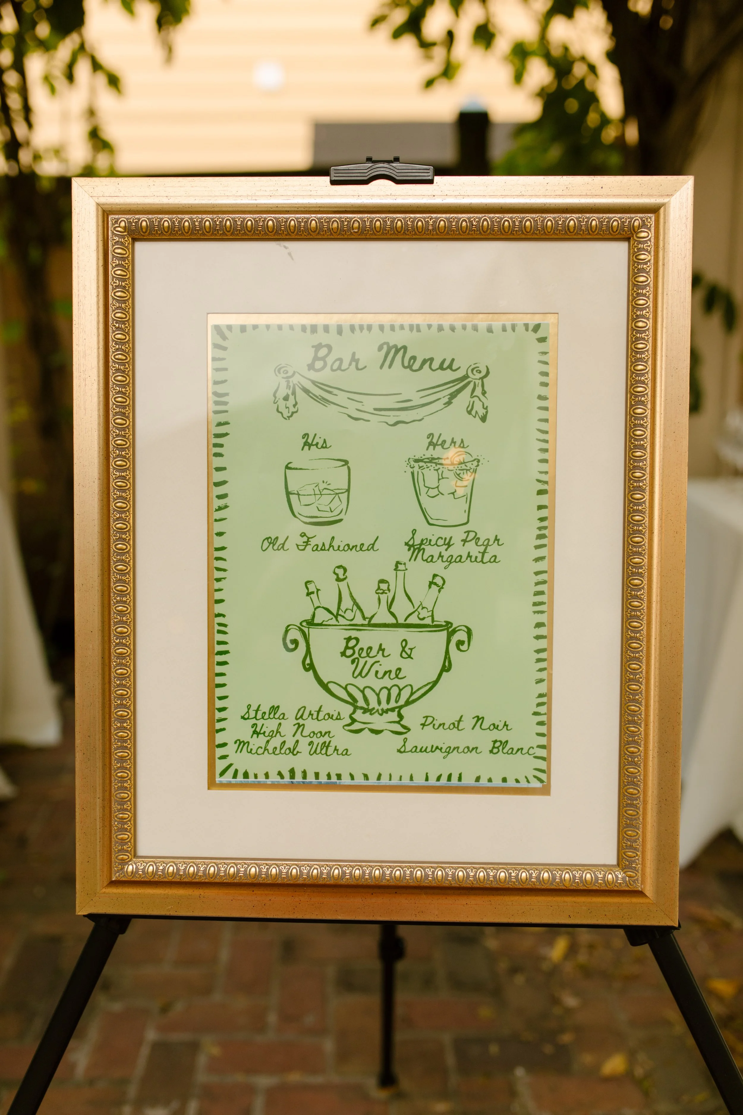 Gold-framed illustrated wedding bar menu displayed outdoors with signature cocktails and a soft green color palette.