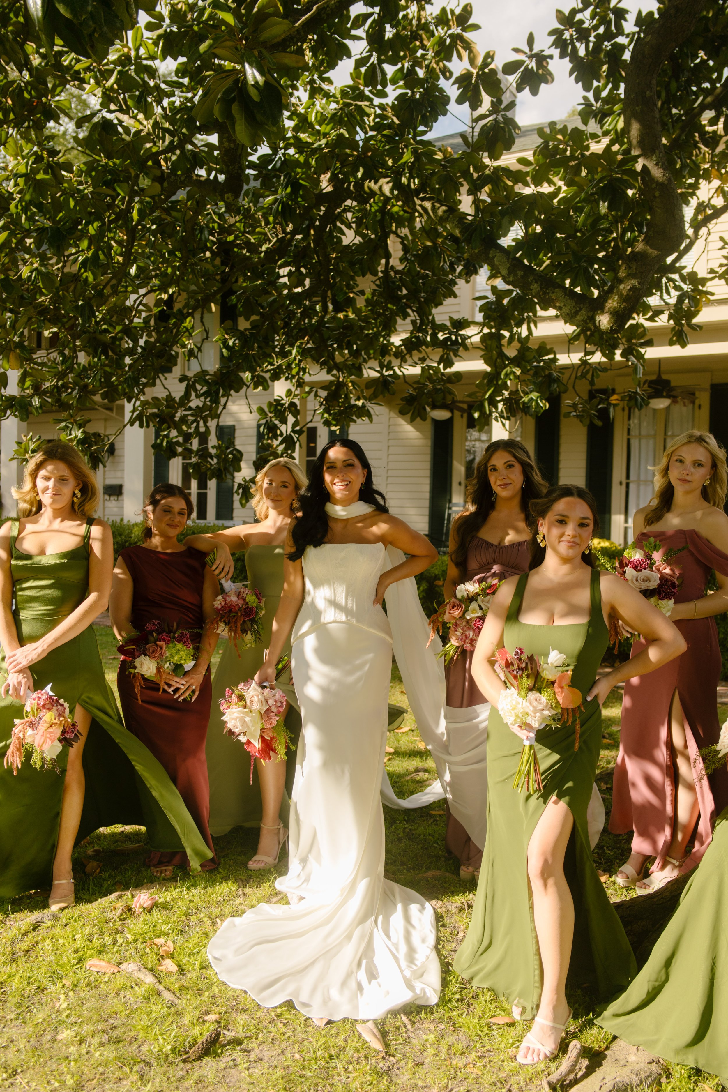 Bride standing with bridesmaids in olive green, burgundy, mauve, and sage dresses, showcasing romantic moody wedding colors.