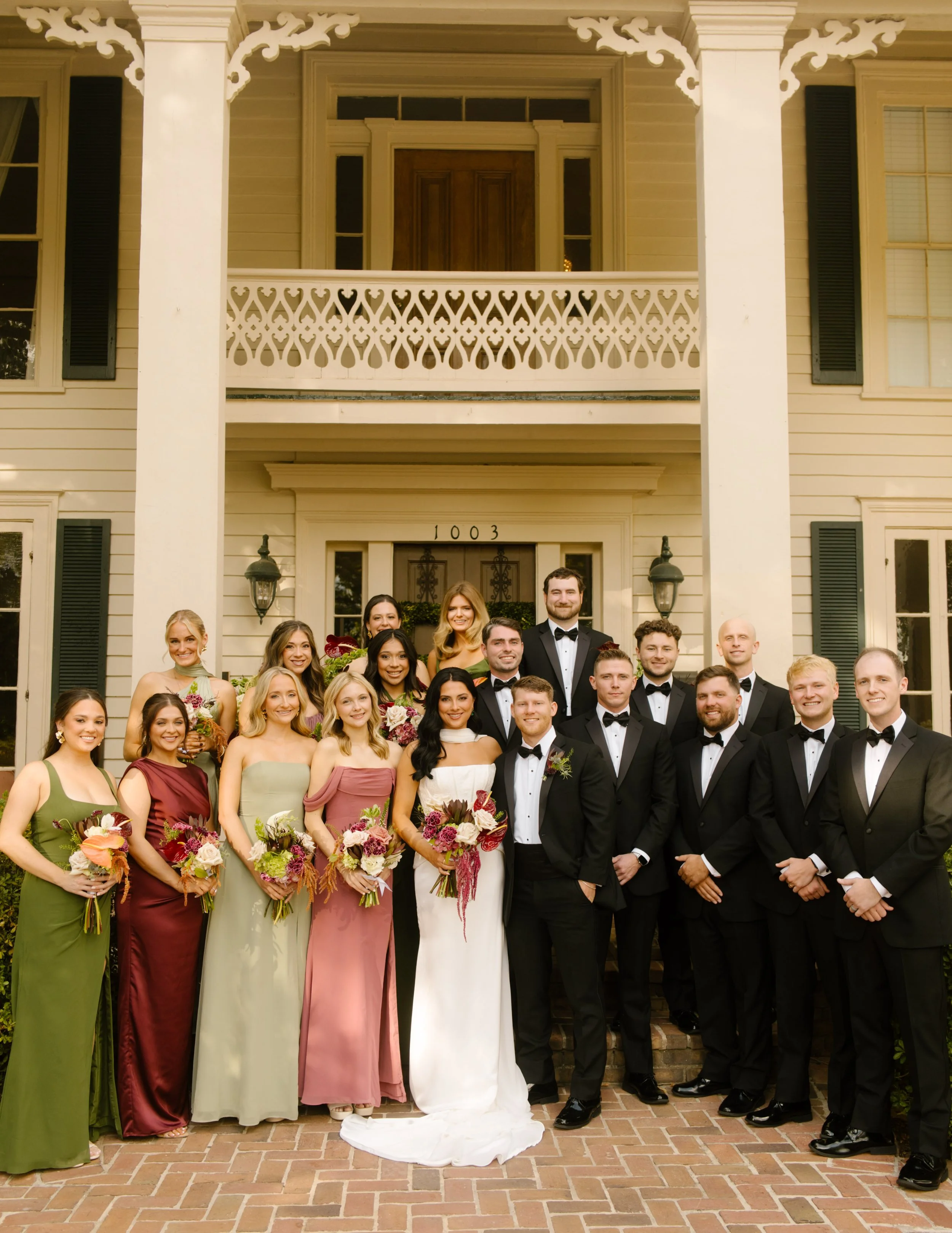 Bride and groom posing with their wedding party outside a historic Southern mansion, with bridesmaids wearing moody wedding colors.