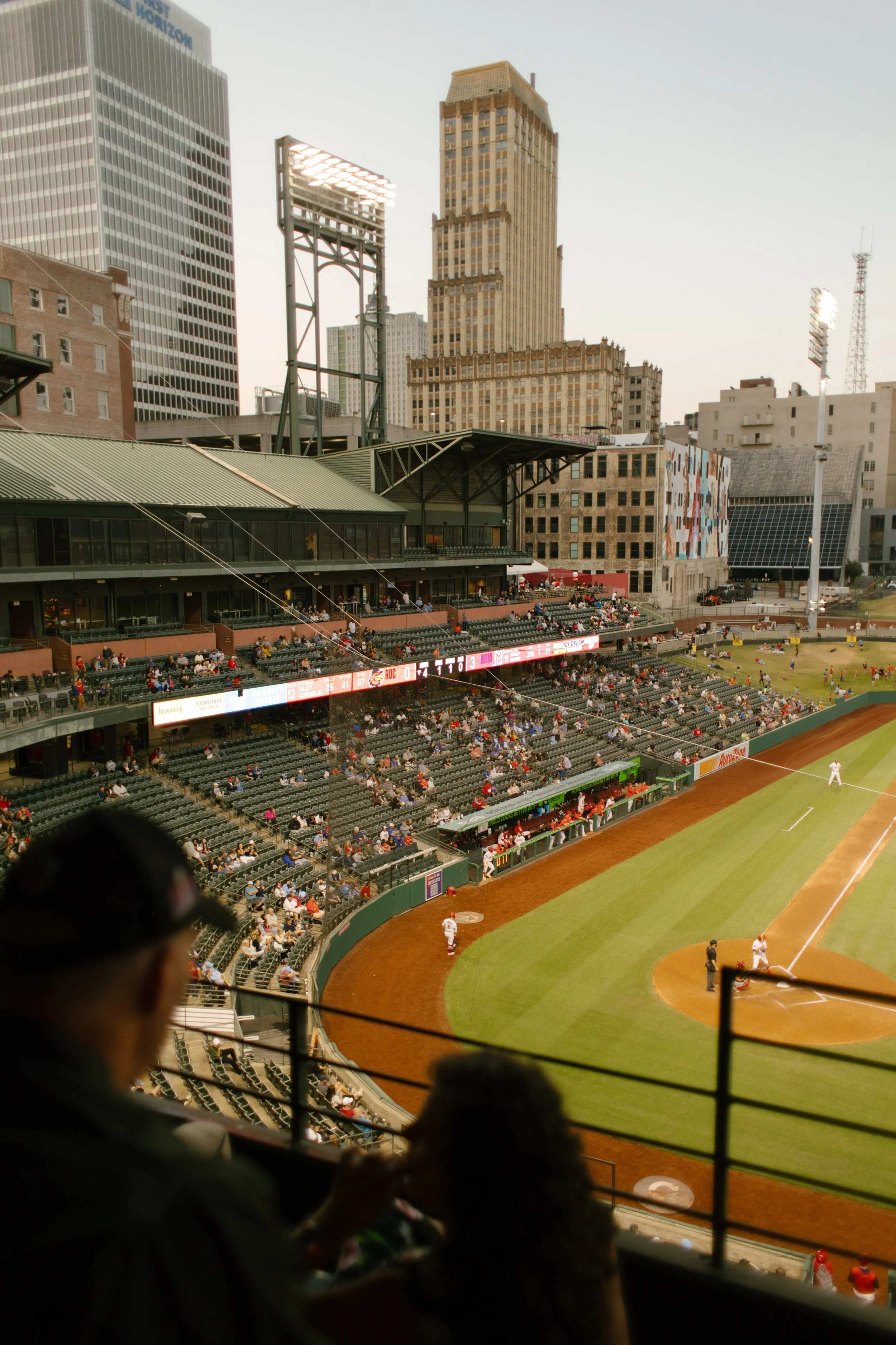 Wide view of a baseball stadium at sunset, with players on the field and the city skyline behind them, inspiring fun engagement photos at a sporting event.