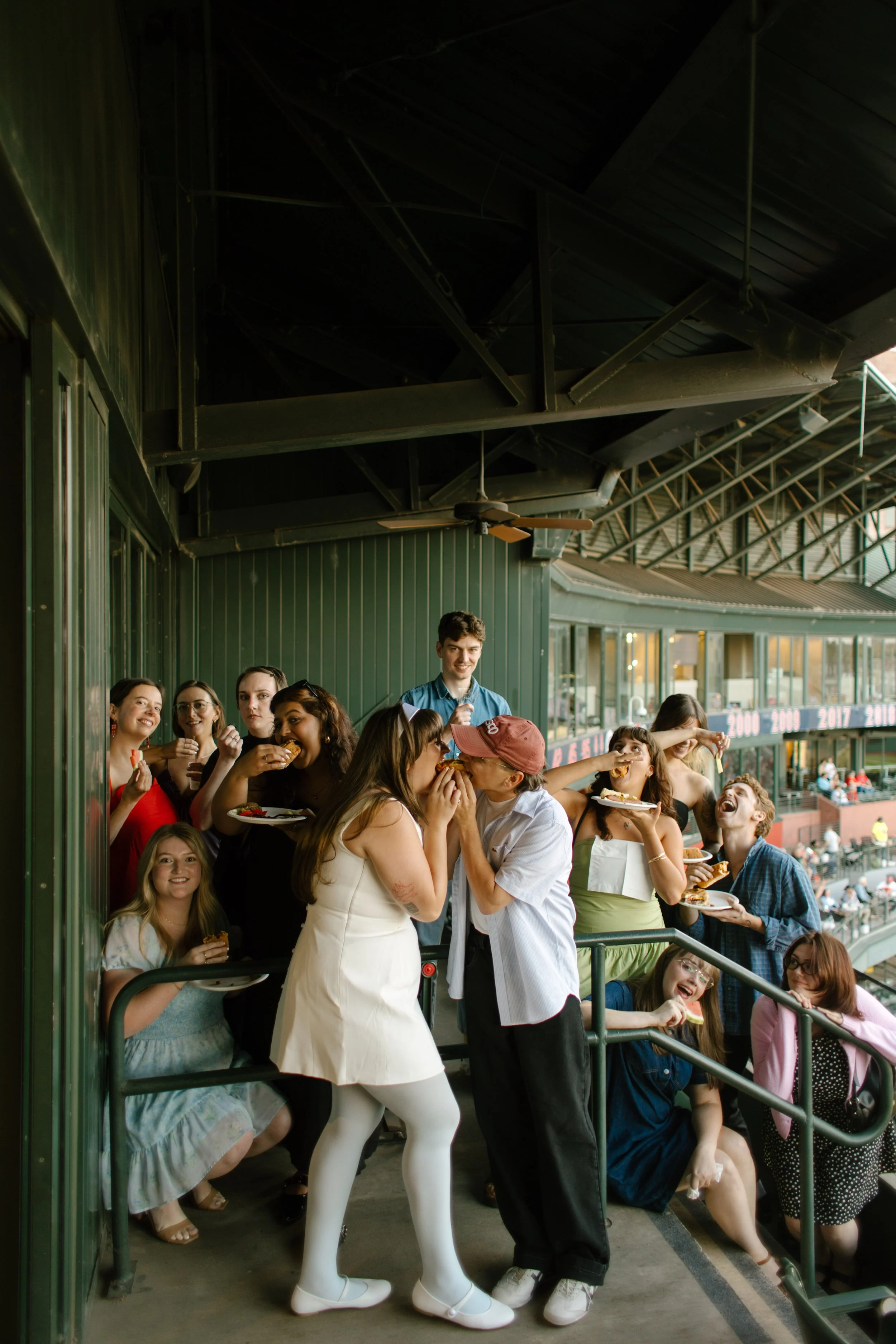 A couple kisses in the middle of a lively group of friends at a baseball stadium, showing the candid energy behind fun engagement photos.