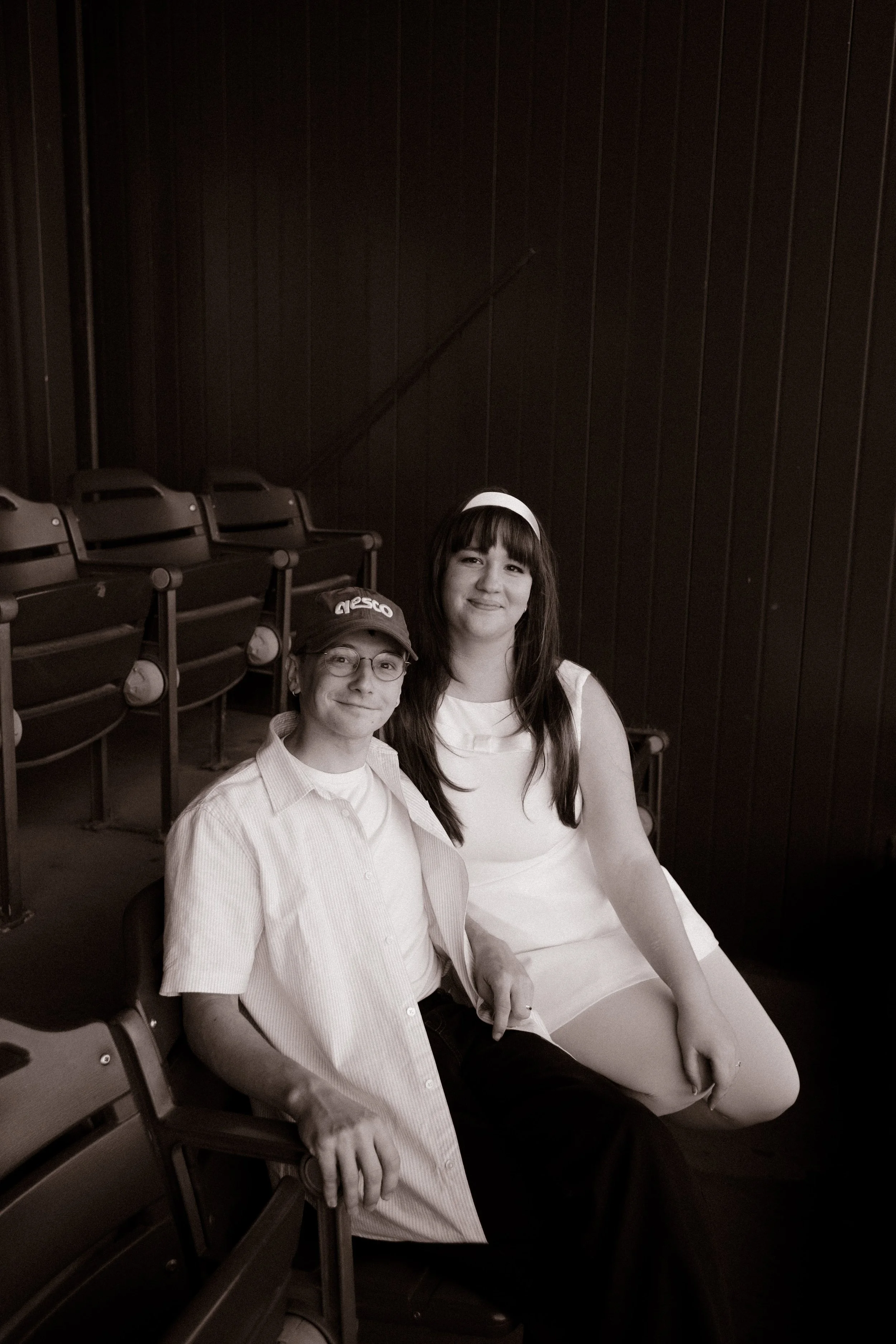Black-and-white portrait of a couple sitting together in stadium seats, smiling at the camera during their baseball-themed celebration.