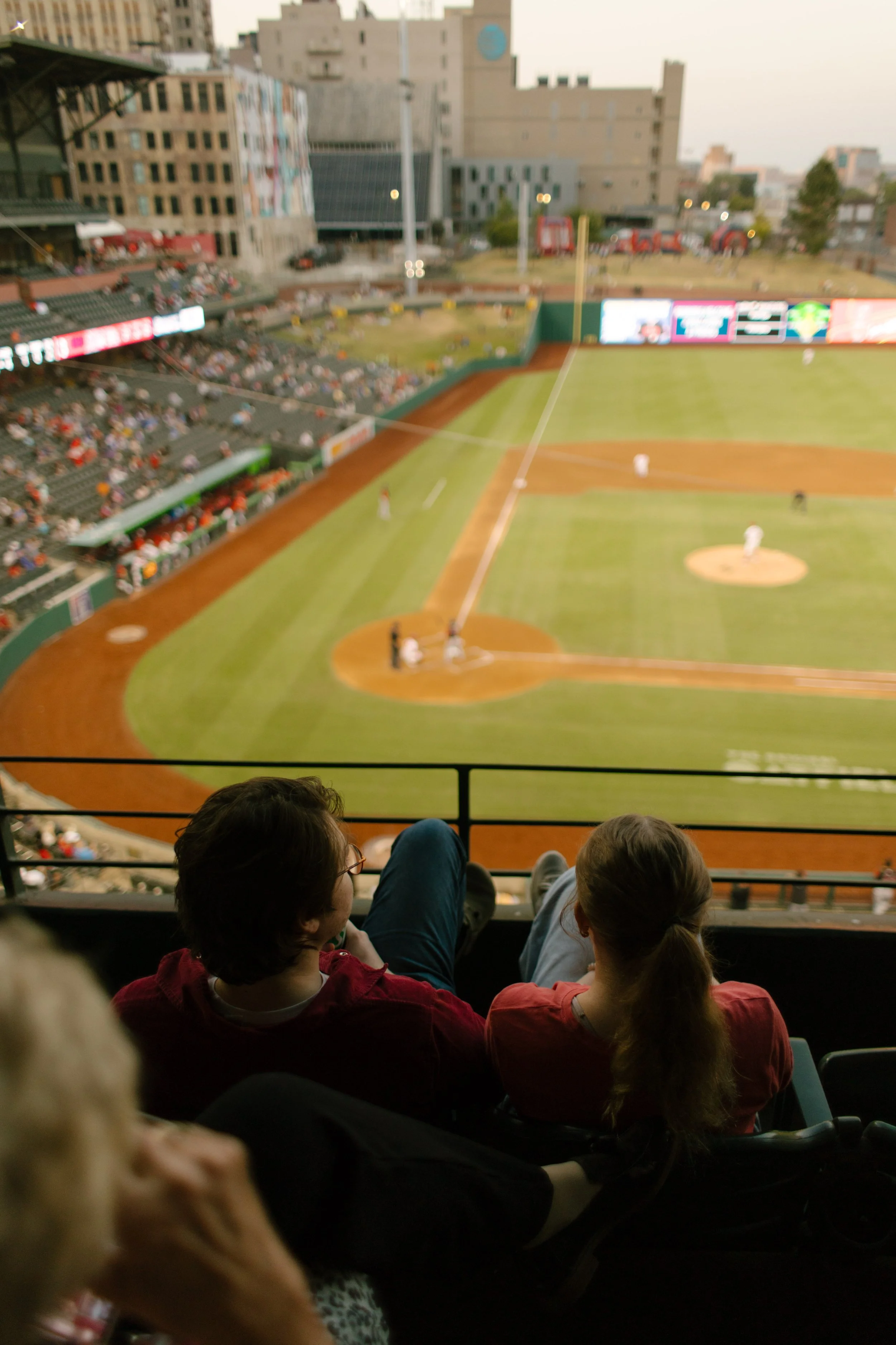 A couple watches a baseball game together from the upper stadium seats, overlooking the diamond and surrounding city skyline.