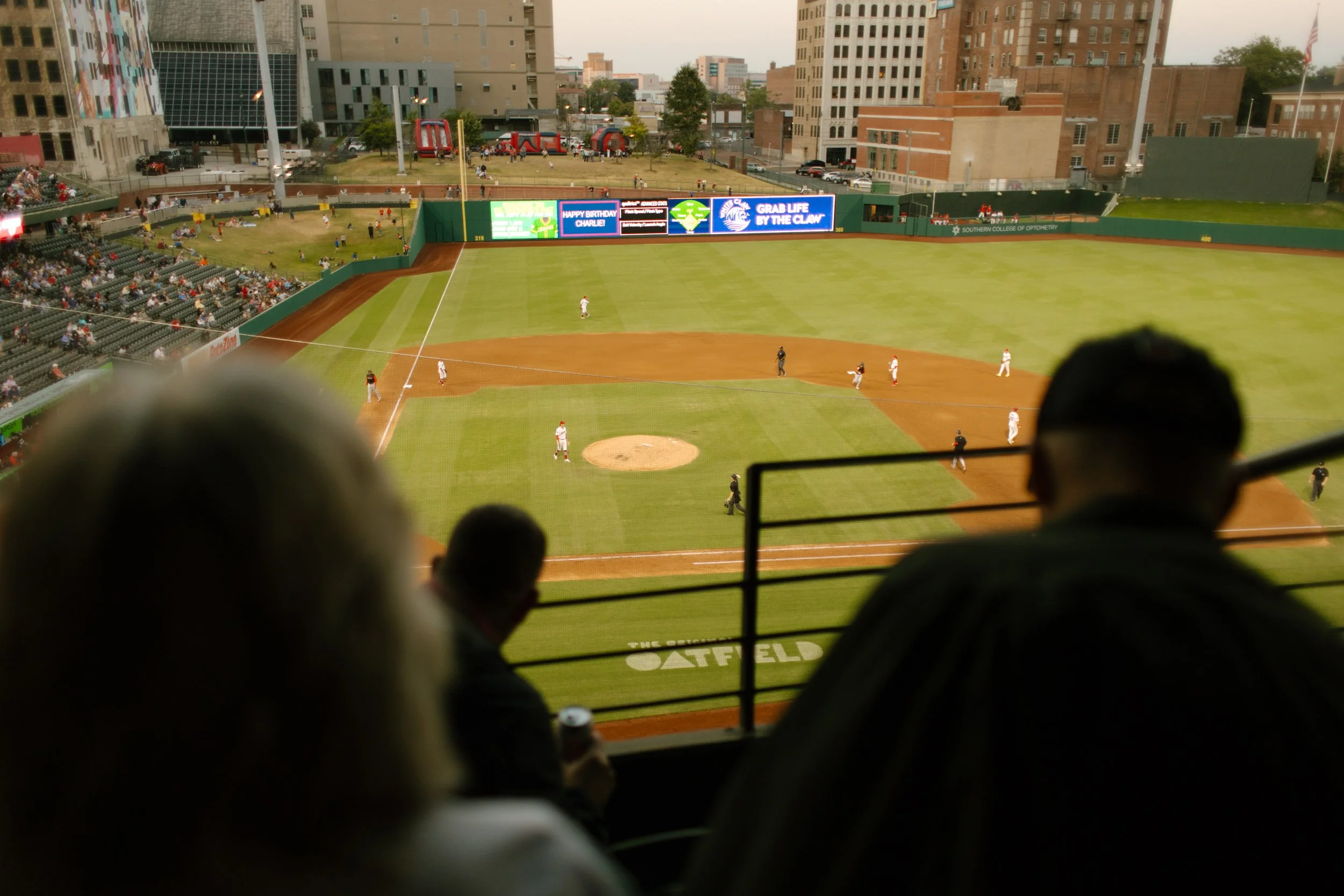 Baseball players on the field during an evening game, photographed from the upper stands with spectators in the foreground.
