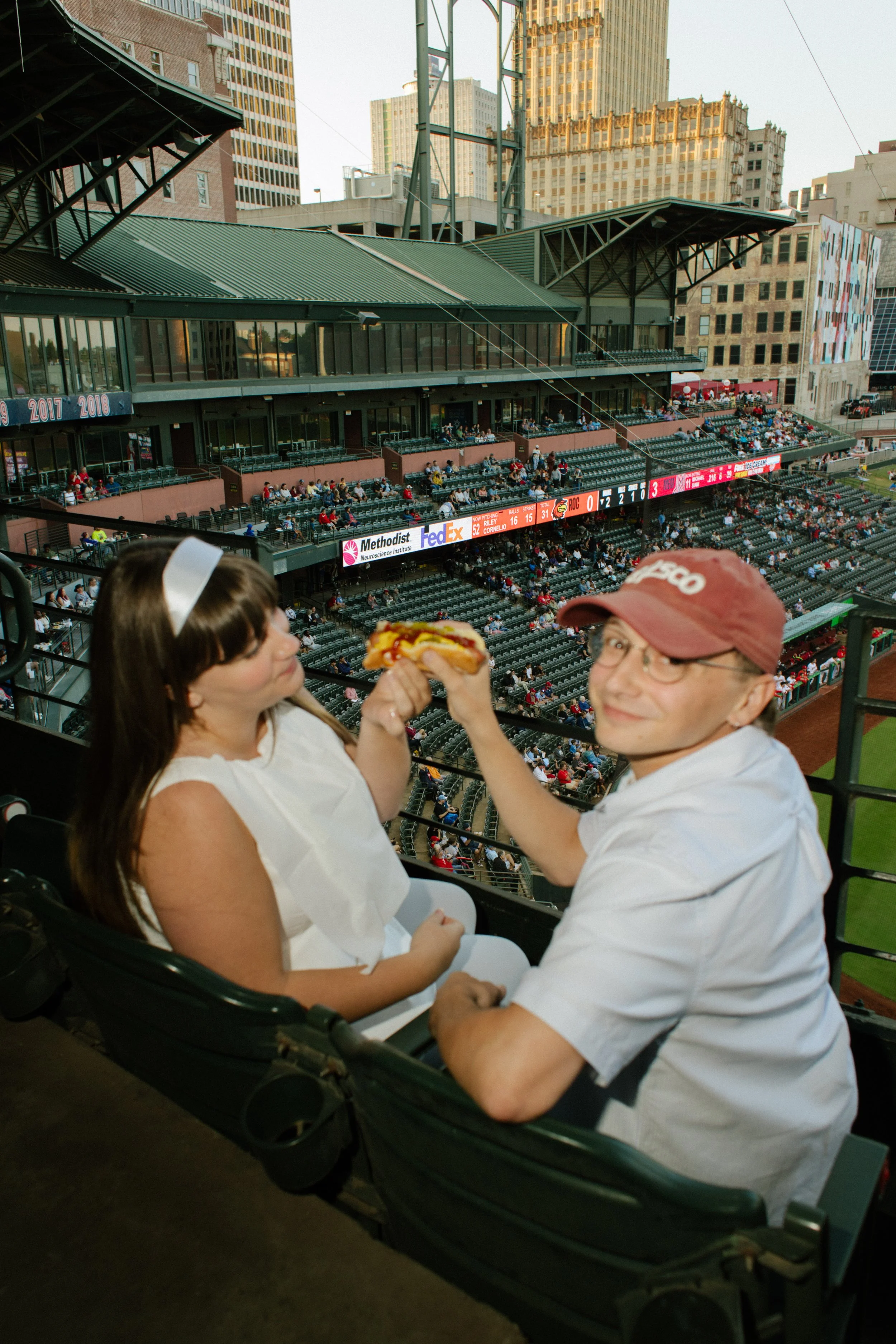 A couple raises a hot dog together while sitting in stadium seats, with the baseball field behind them, offering inspiration for fun engagement photos.