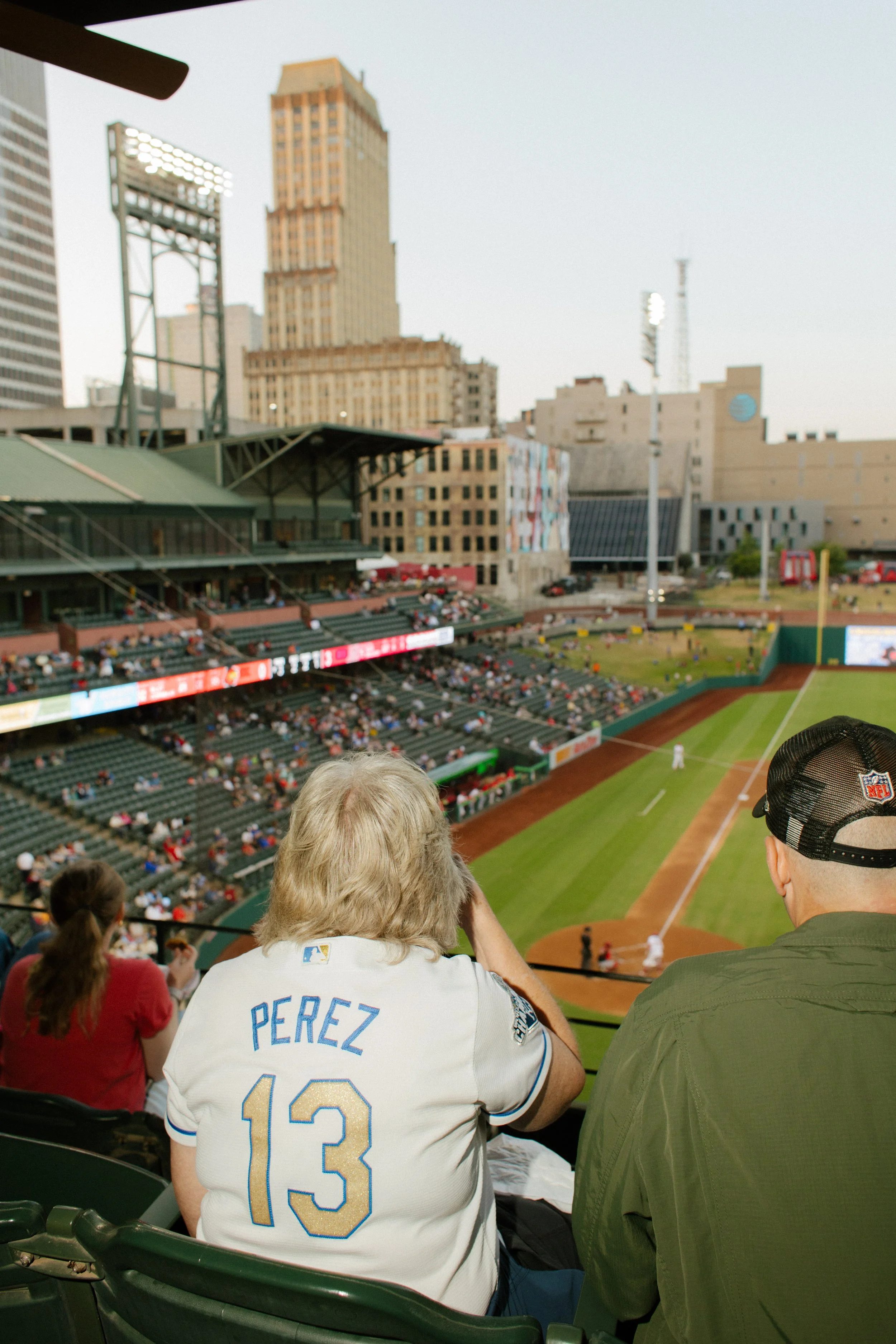 Baseball fans watch the game from elevated seats, including a spectator wearing a Perez number 13 jersey, with the field and city skyline beyond.