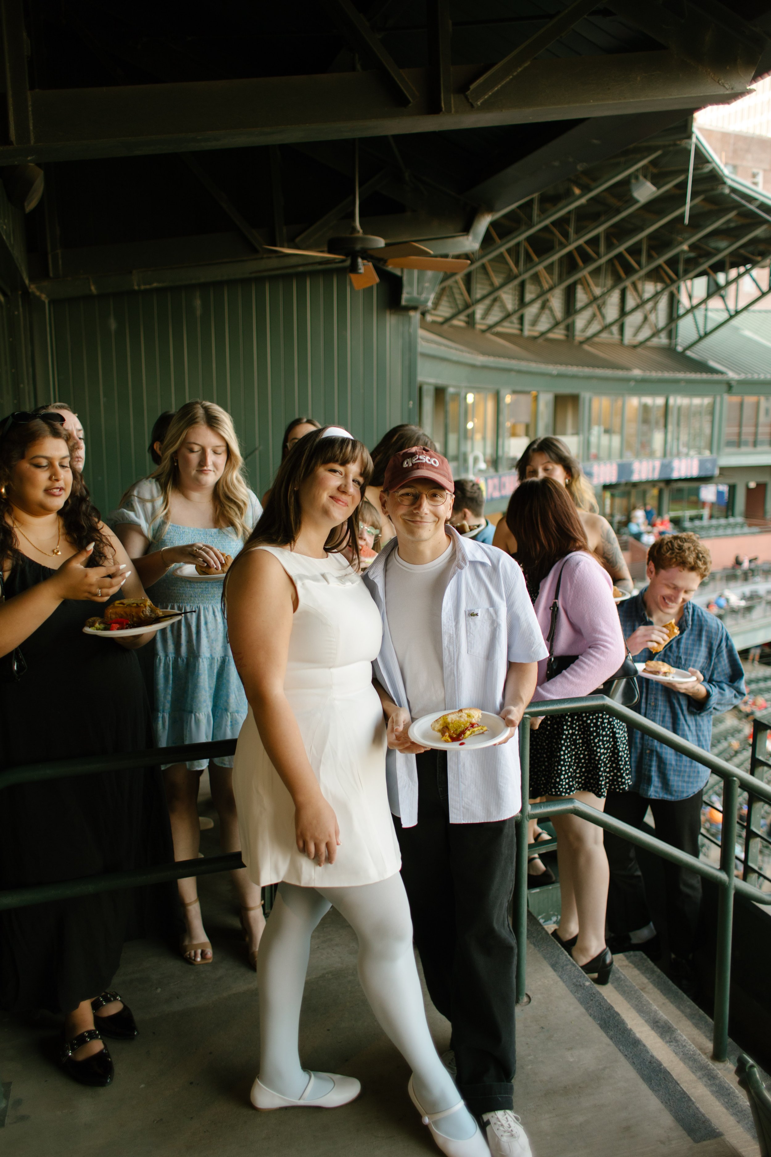 A couple poses together on a stadium balcony while friends enjoy food and drinks around them during a baseball-themed rehearsal dinner.