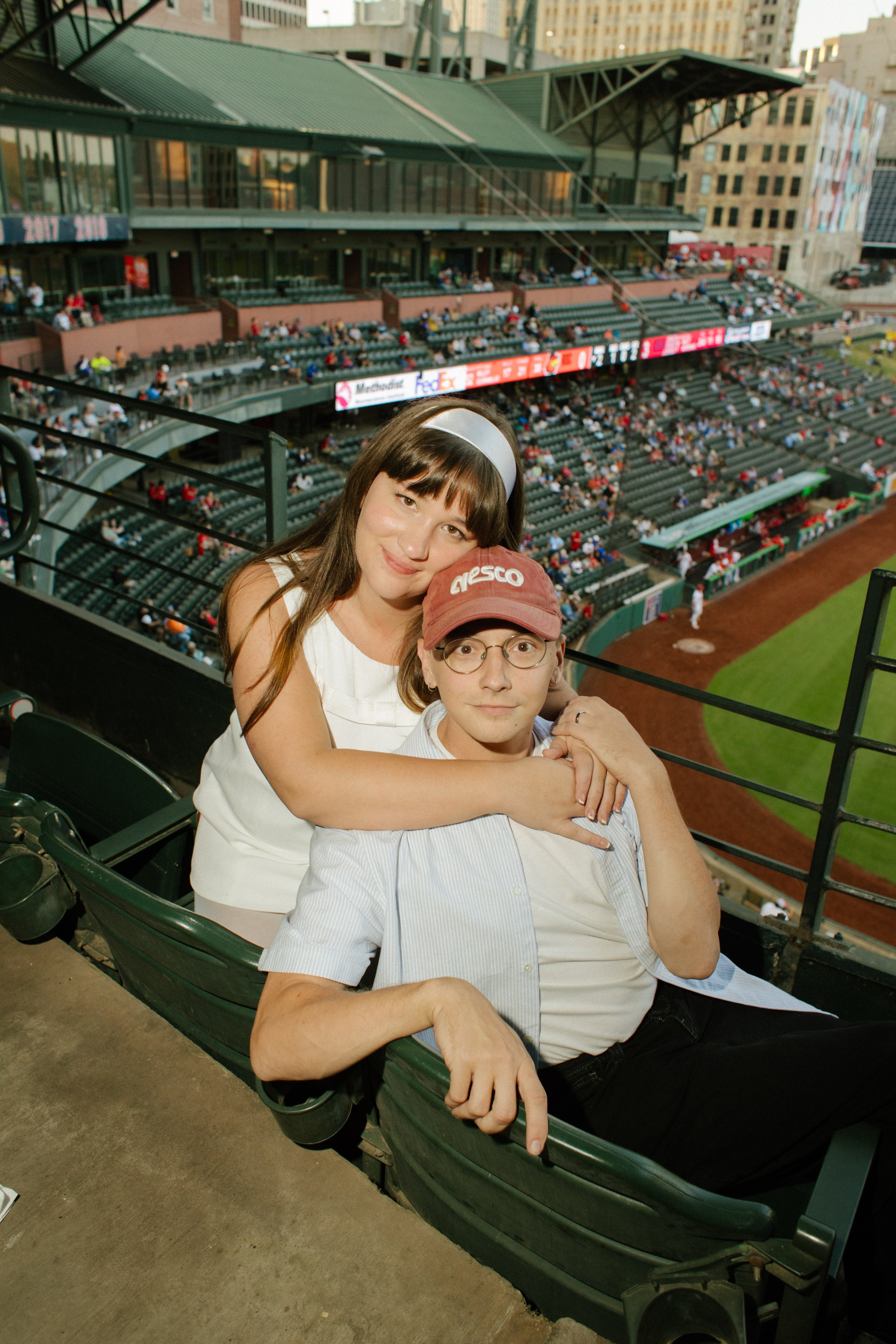 A couple cuddles together in baseball stadium seats overlooking the field, showing the relaxed, playful moments that inspire fun engagement photos.