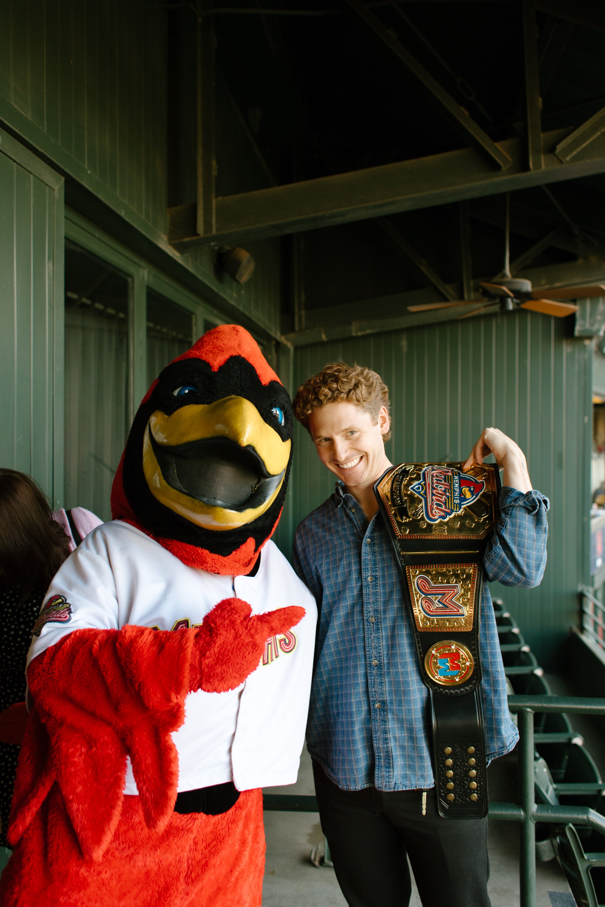A smiling guest poses with a red bird baseball mascot and a championship-style belt on the stadium balcony.