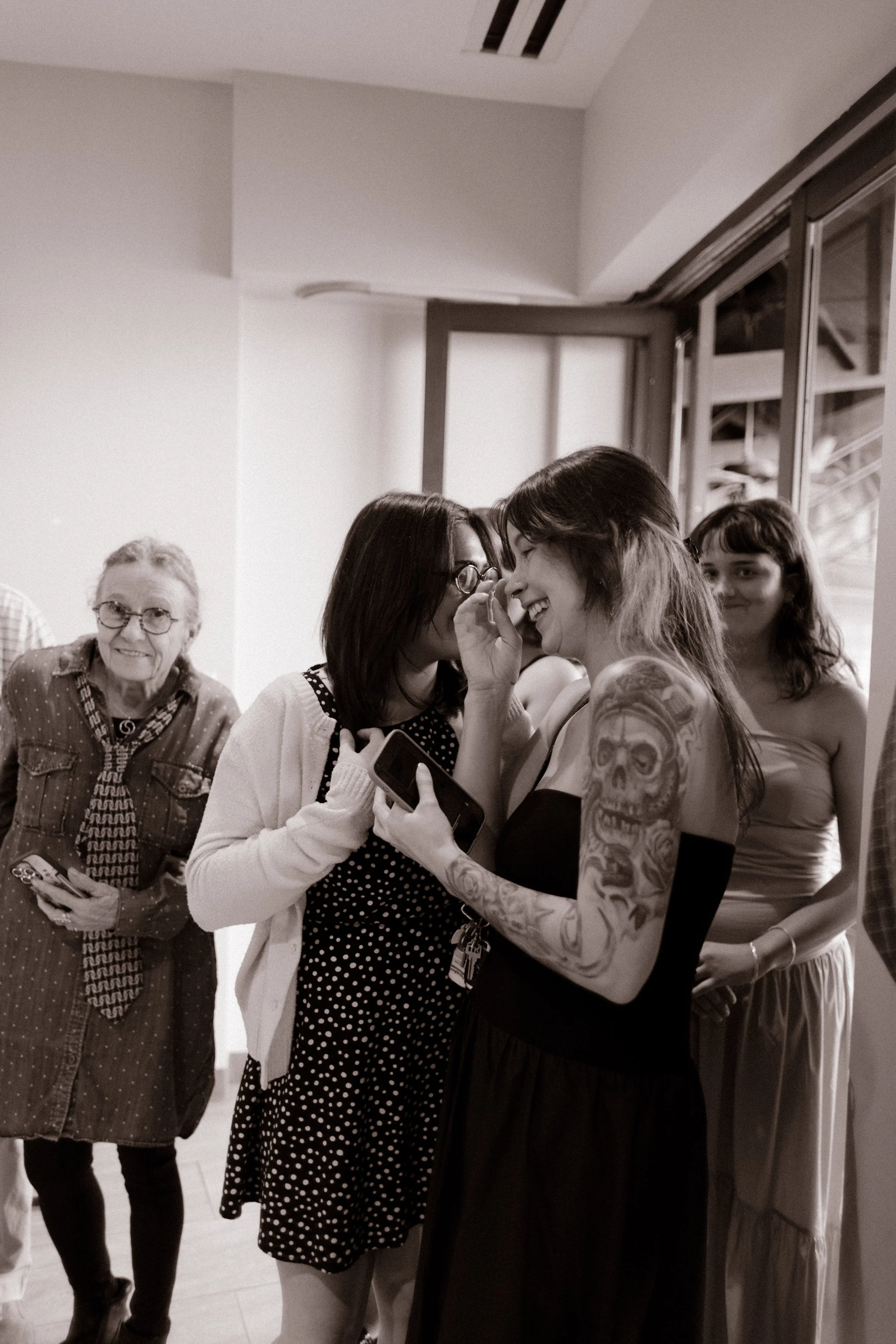 Black-and-white photograph of two women sharing a laugh during a baseball-themed rehearsal dinner, with other guests gathered nearby.