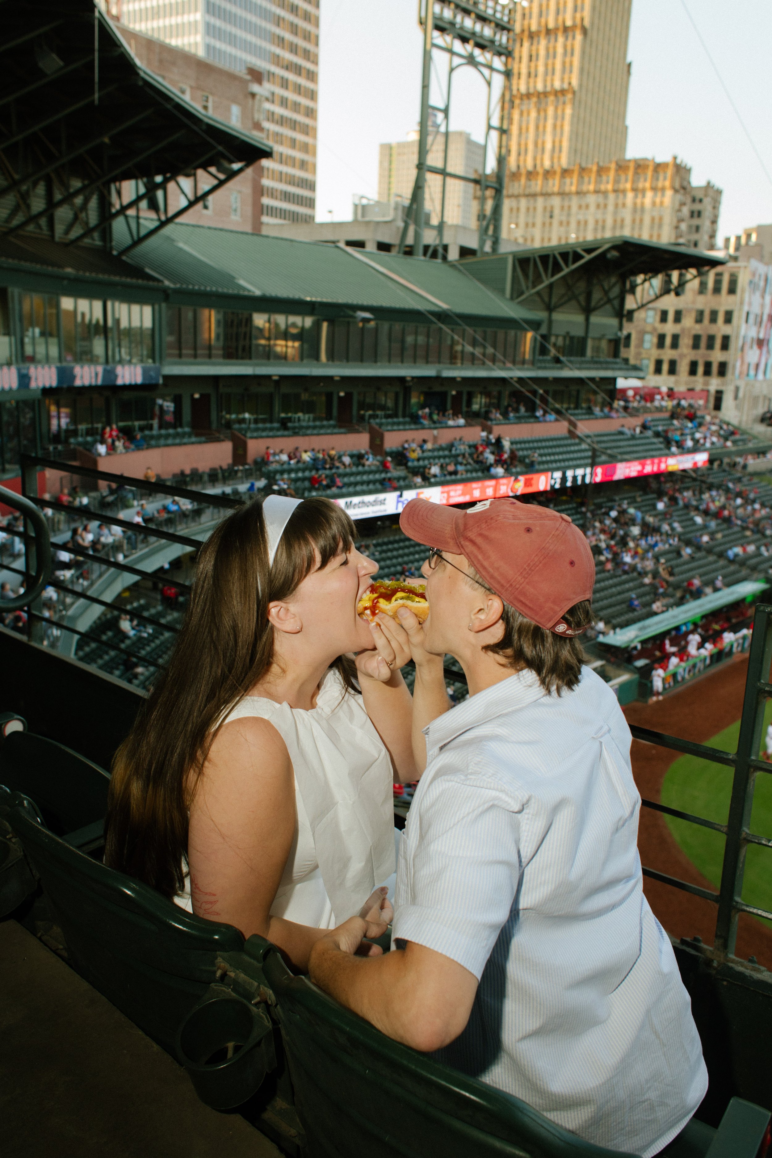 A couple playfully takes bites from the same hot dog in stadium seats, inspiring fun engagement photos centered around a favorite activity.
