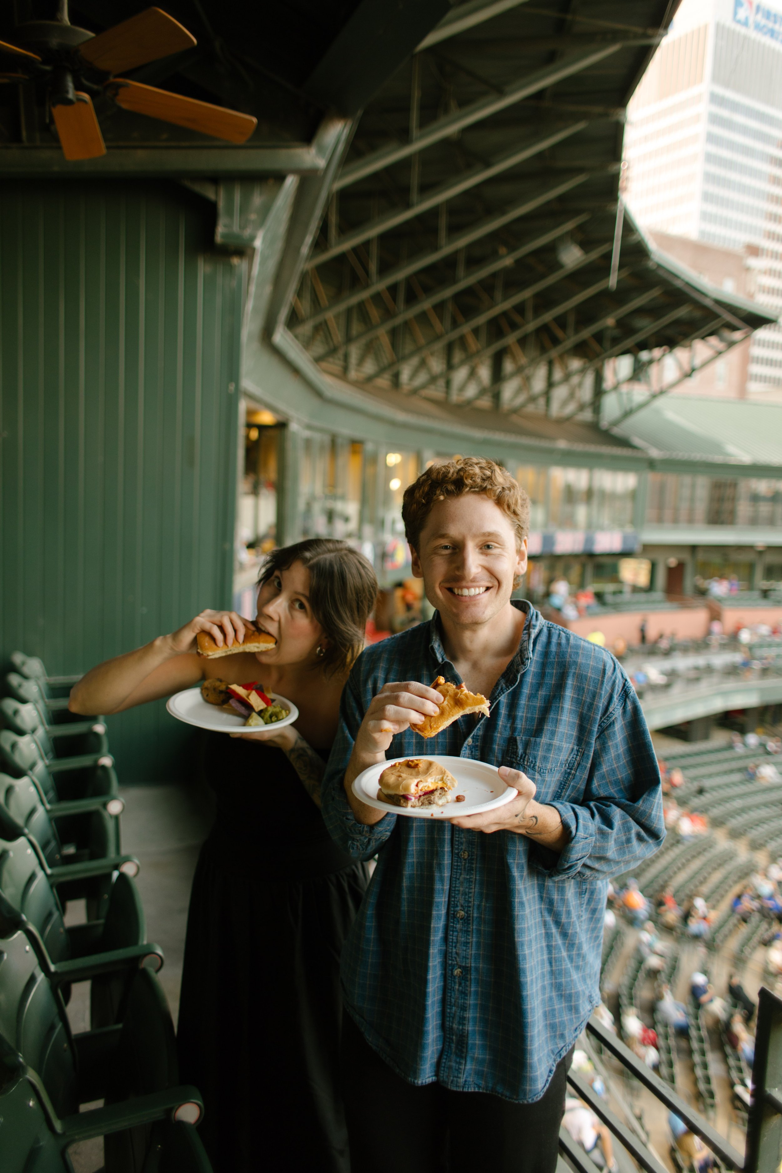 Two guests enjoy stadium food on a balcony overlooking the baseball field, offering candid inspiration for fun engagement photos.