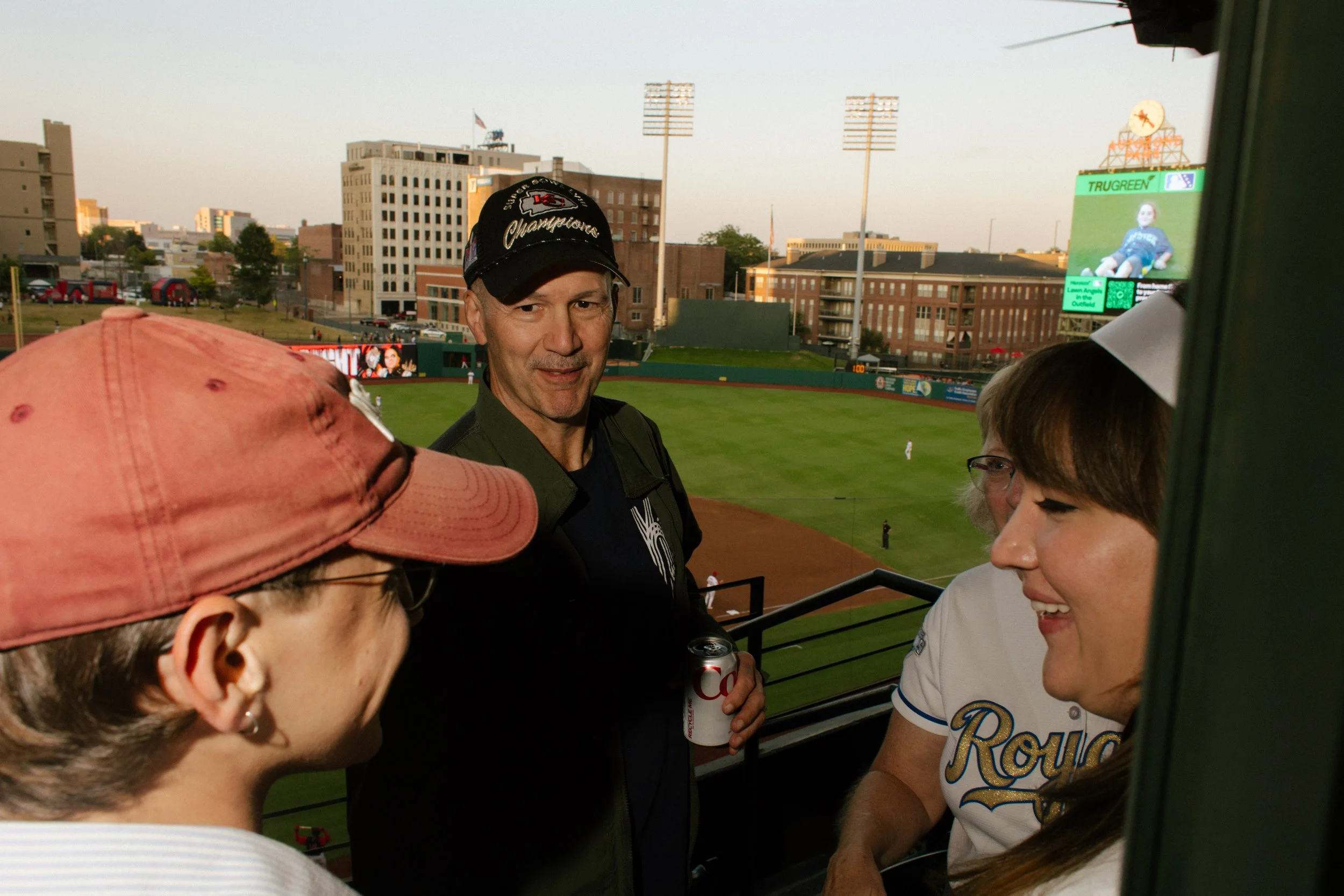 Three guests chat near a baseball stadium balcony at sunset, with the field and illuminated scoreboard in the background.