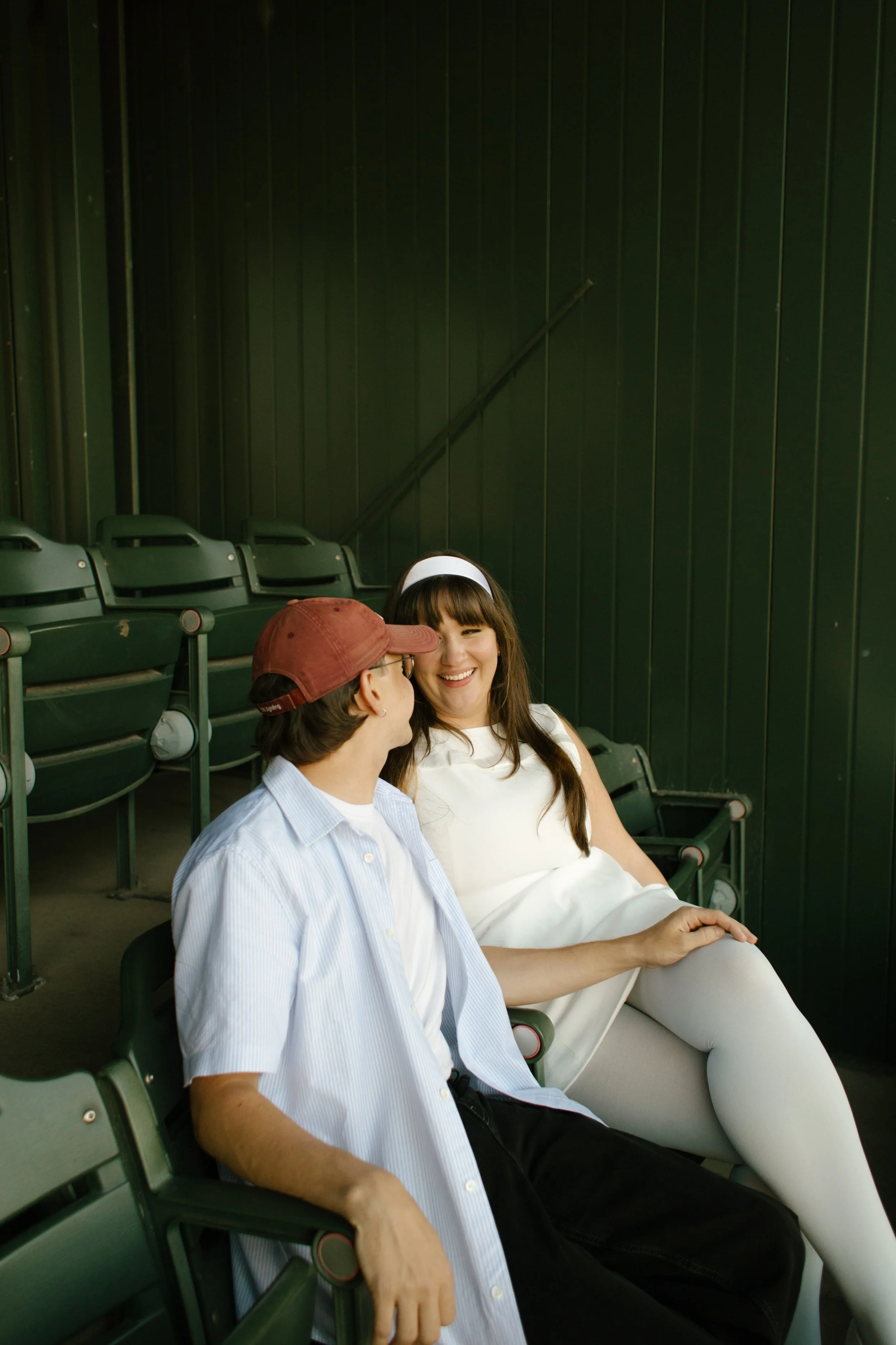 A couple laughs together while sitting in a quiet section of green stadium seats during their baseball-themed rehearsal dinner.