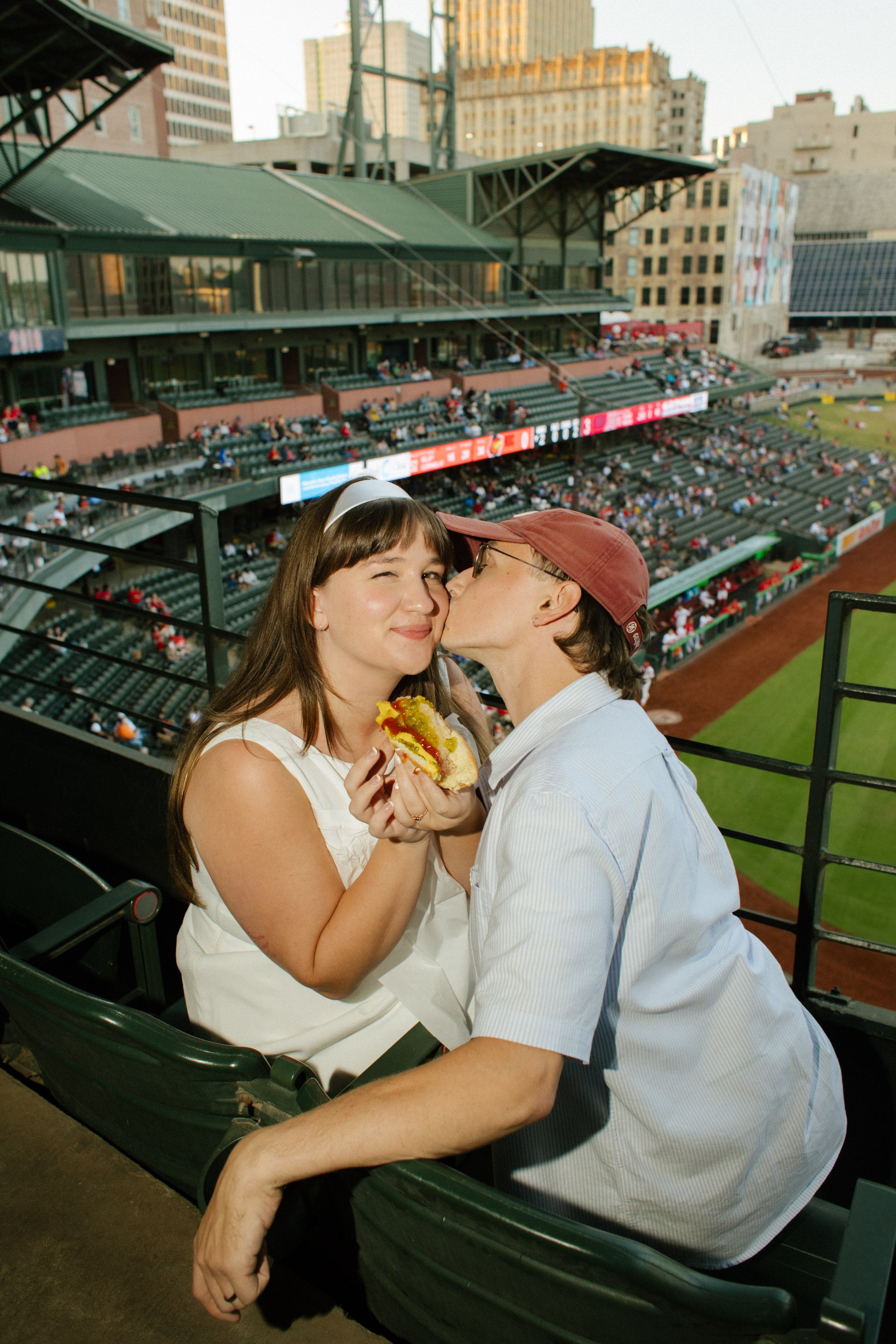 A woman holding a hot dog receives a kiss on the cheek from her partner in baseball stadium seats, inspiring fun engagement photos centered on shared activities.