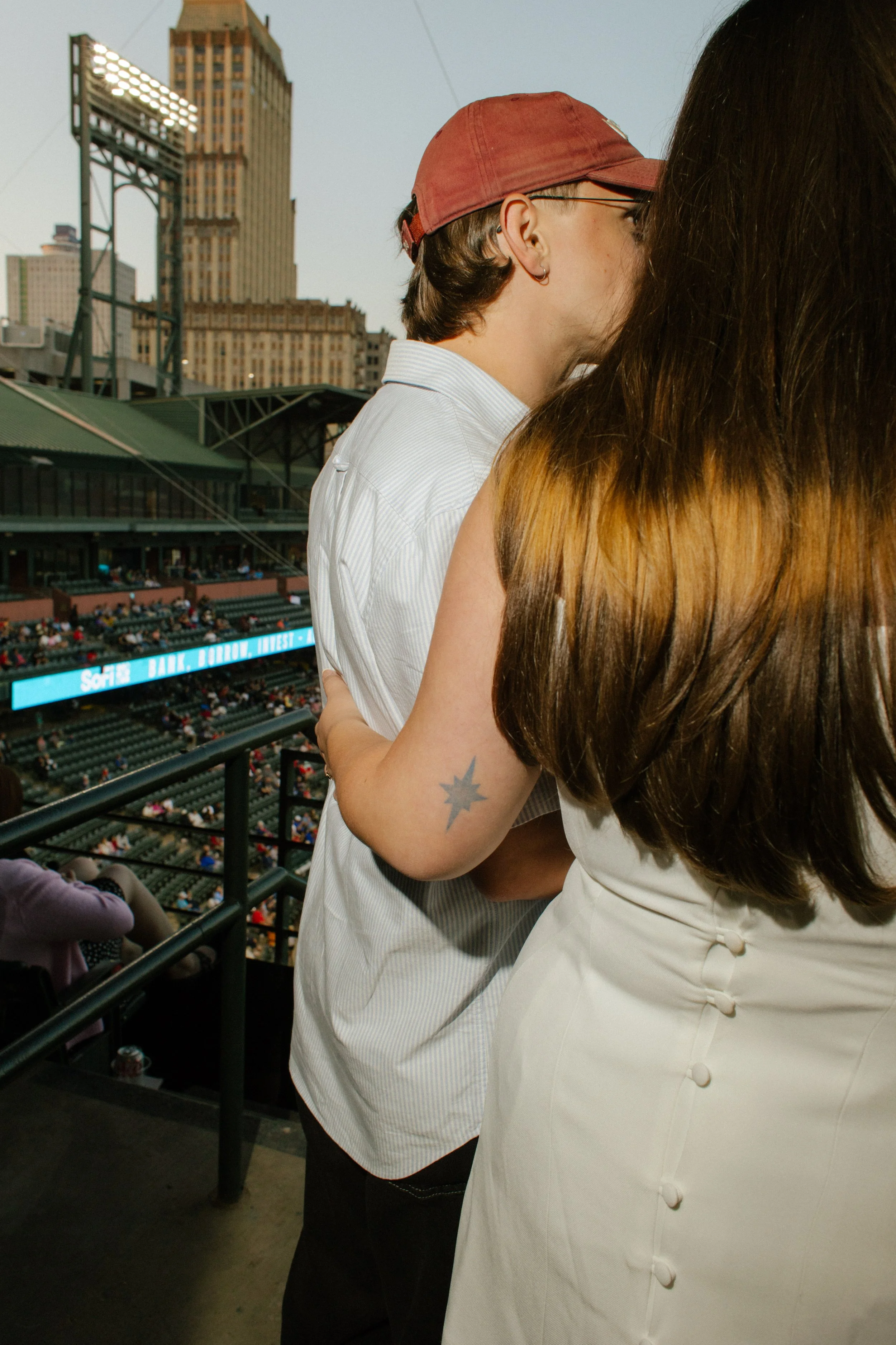 A couple embraces beside the railing overlooking a baseball stadium, showing how everyday experiences can inspire fun engagement photos.