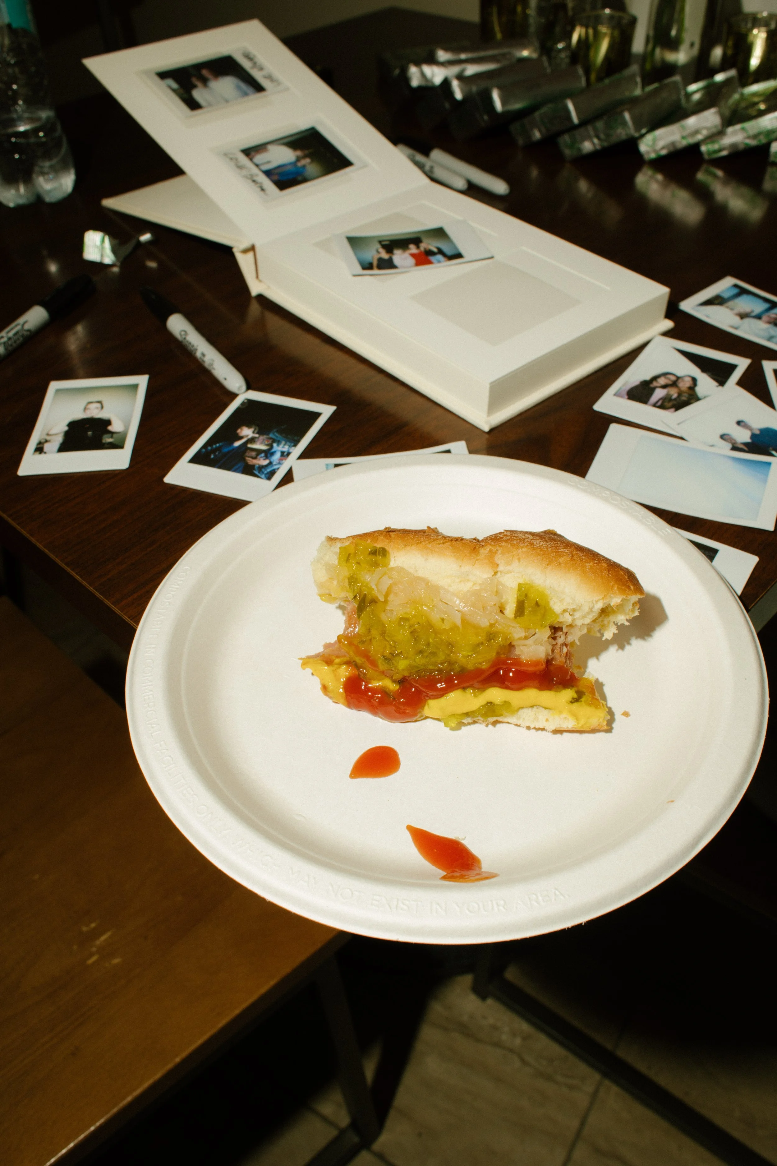 Close-up of a partially eaten stadium sandwich on a white plate surrounded by instant photographs and an open photo album.
