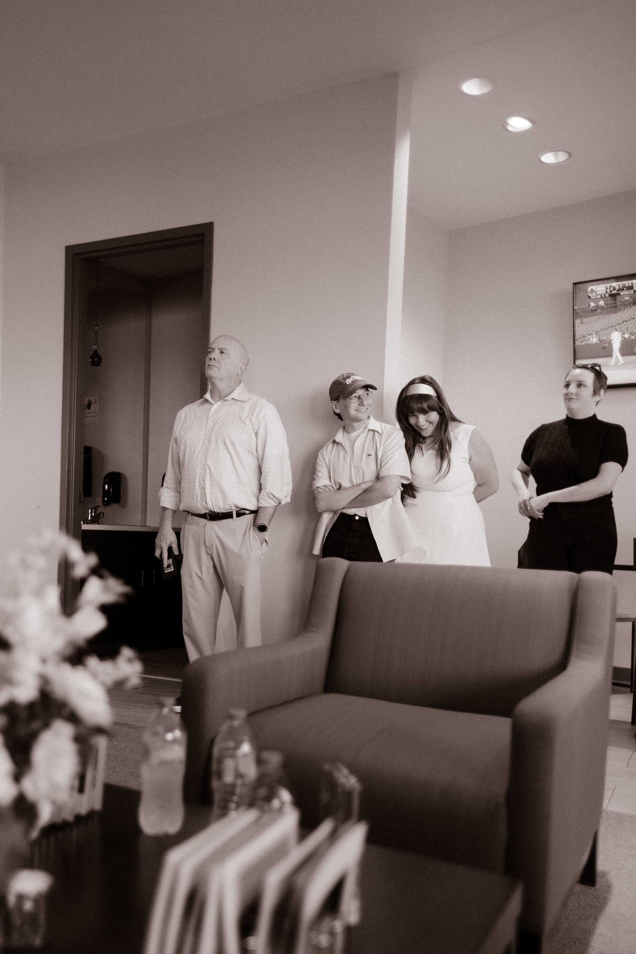 Black-and-white candid photograph of wedding rehearsal guests standing together in a stadium lounge, with a couch and framed baseball photograph nearby.