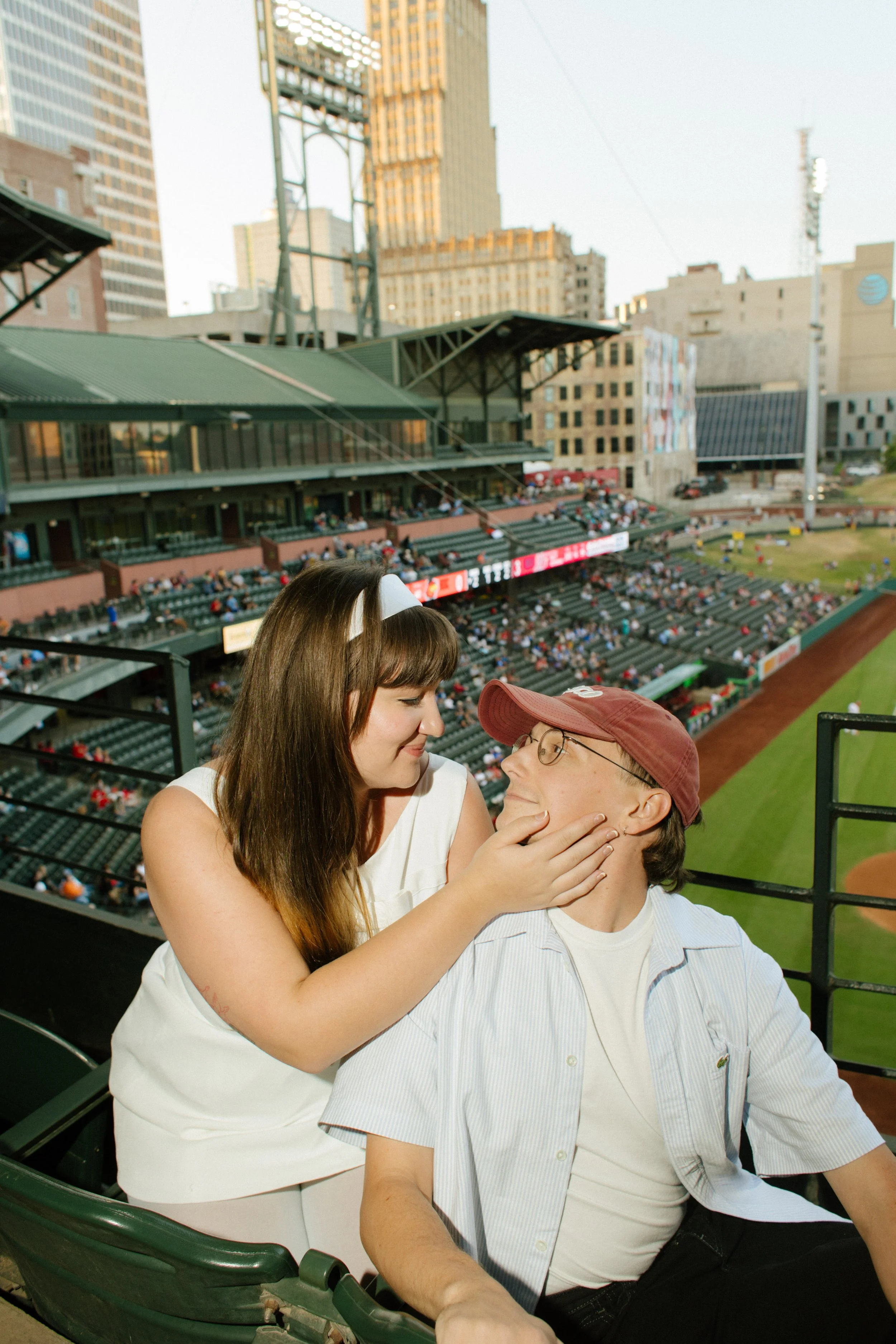A woman in a white dress gently holds her partner's face as they smile at each other in baseball stadium seats overlooking the field.