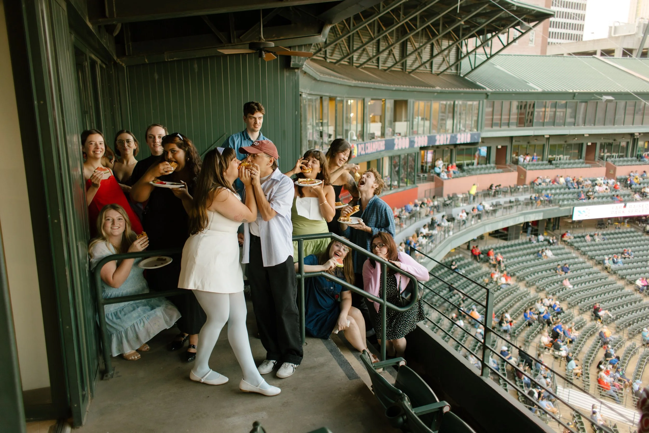 A couple kisses on a stadium balcony while friends gather around them with food and drinks, capturing the playful energy that makes fun engagement photos memorable.
