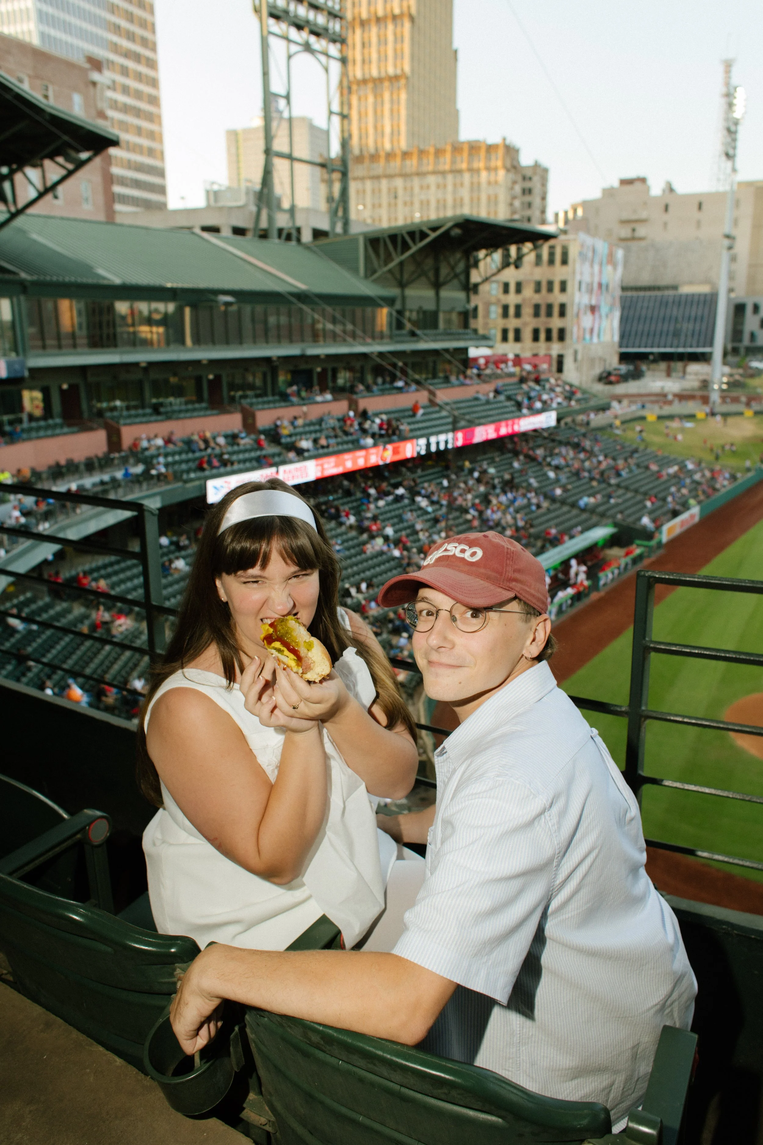 A woman in a white dress takes a bite of a hot dog while her partner smiles beside her, showing the candid moments that inspire fun engagement photos.