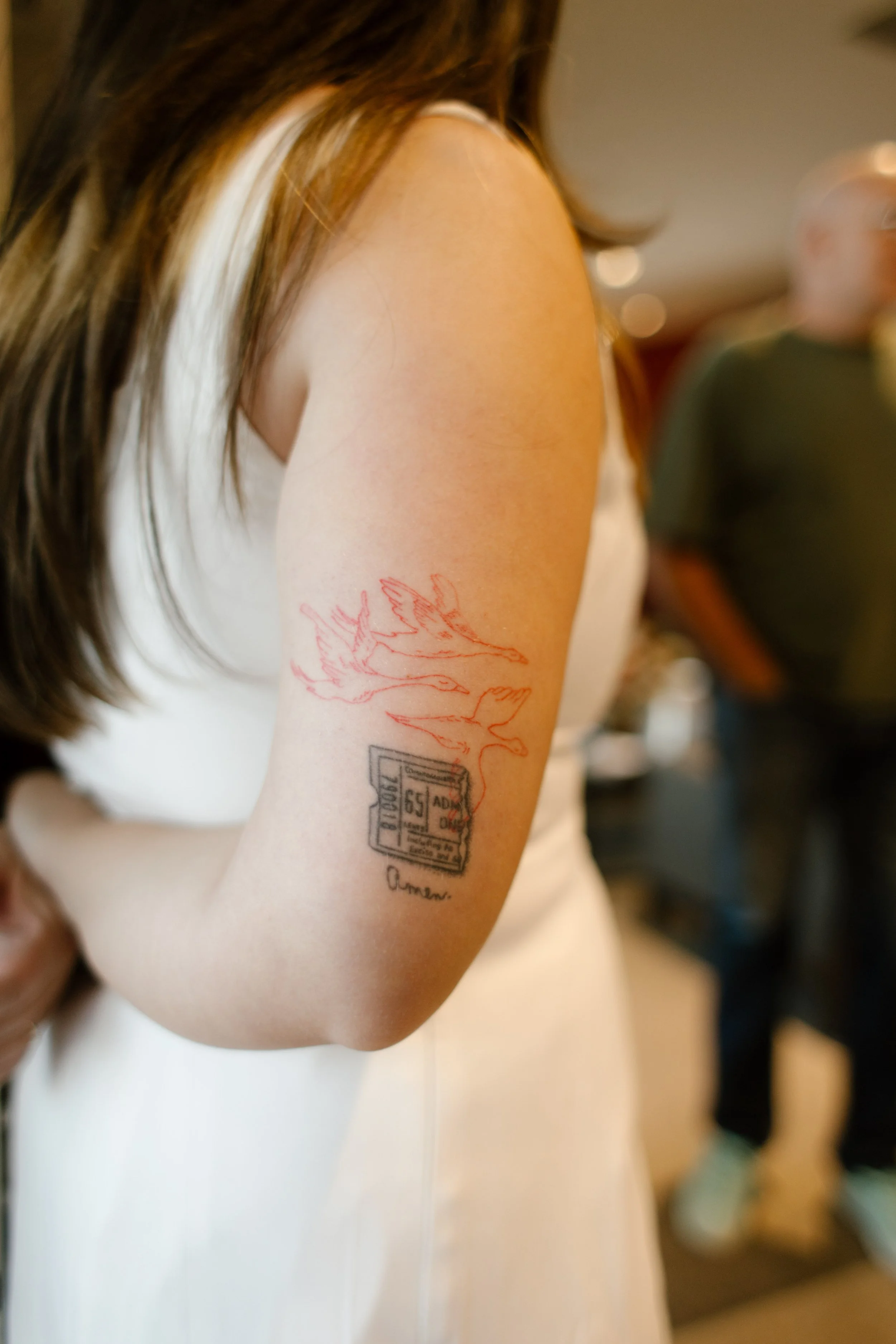 Close-up of a woman's fine-line arm tattoo featuring red artwork and a small ticket design, photographed during a baseball-themed rehearsal dinner.