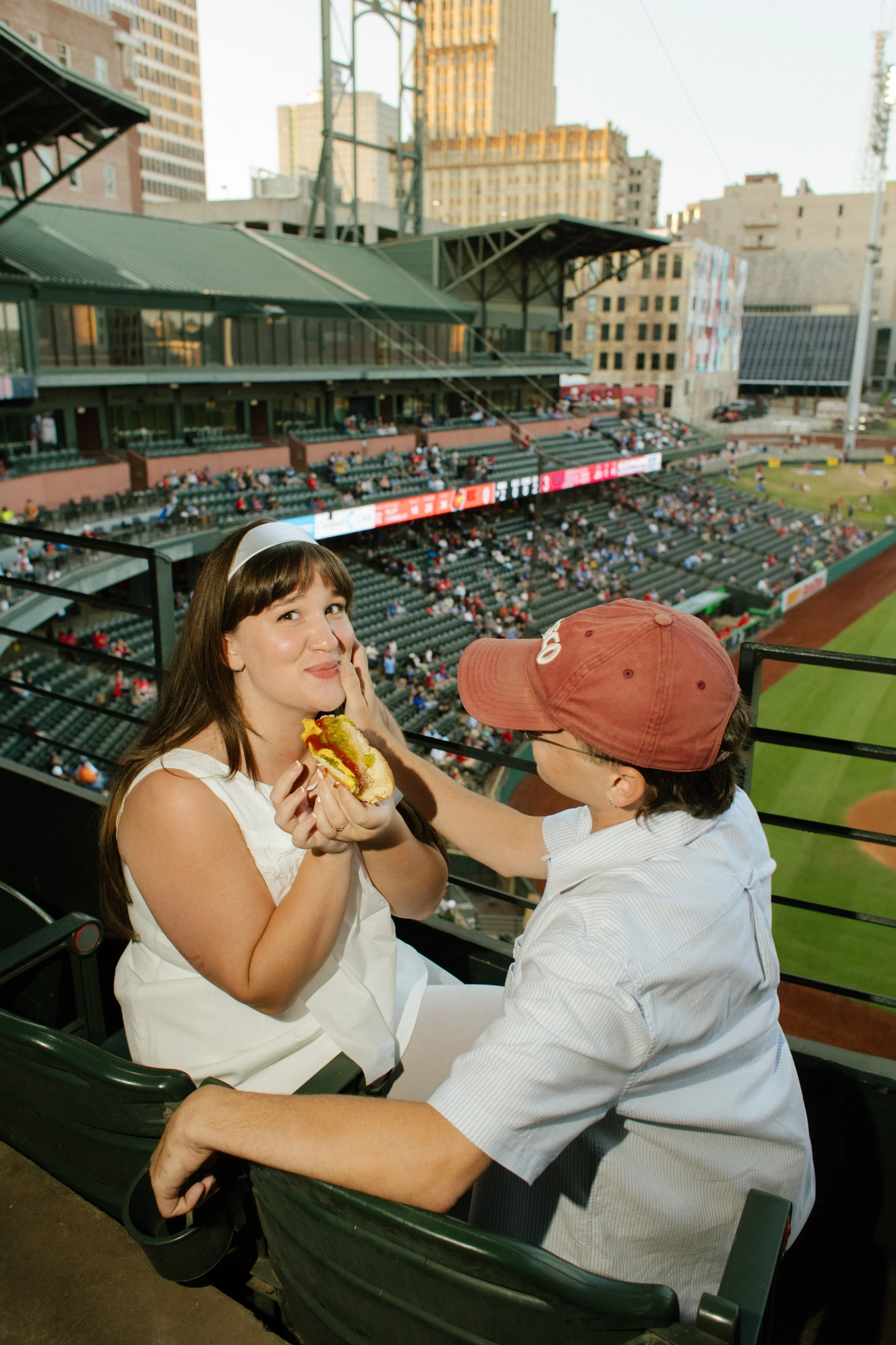 A couple shares a playful moment over a hot dog in their stadium seats, offering inspiration for fun engagement photos centered on favorite activities.