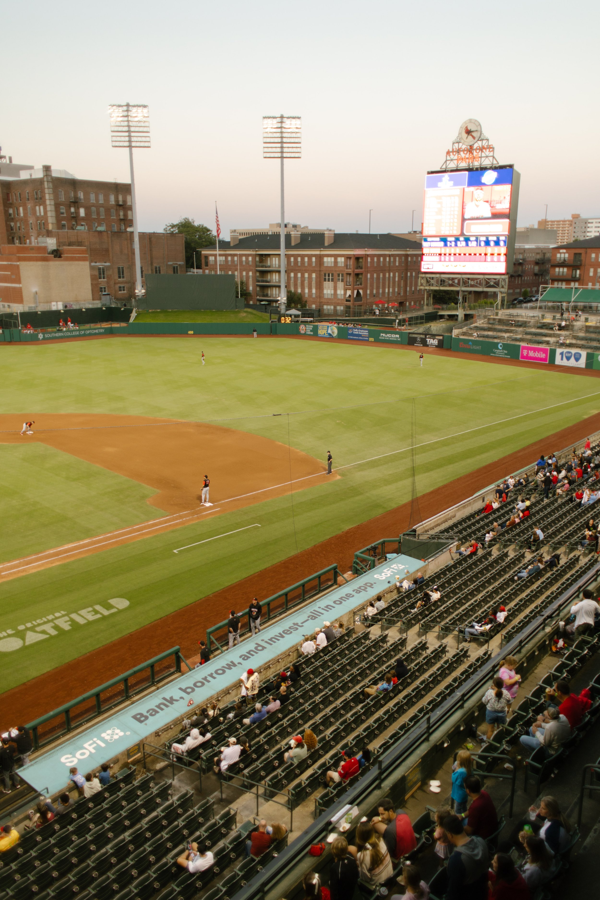 Wide photograph of a baseball stadium at dusk, with players on the field, spectators in the stands, and an illuminated scoreboard.