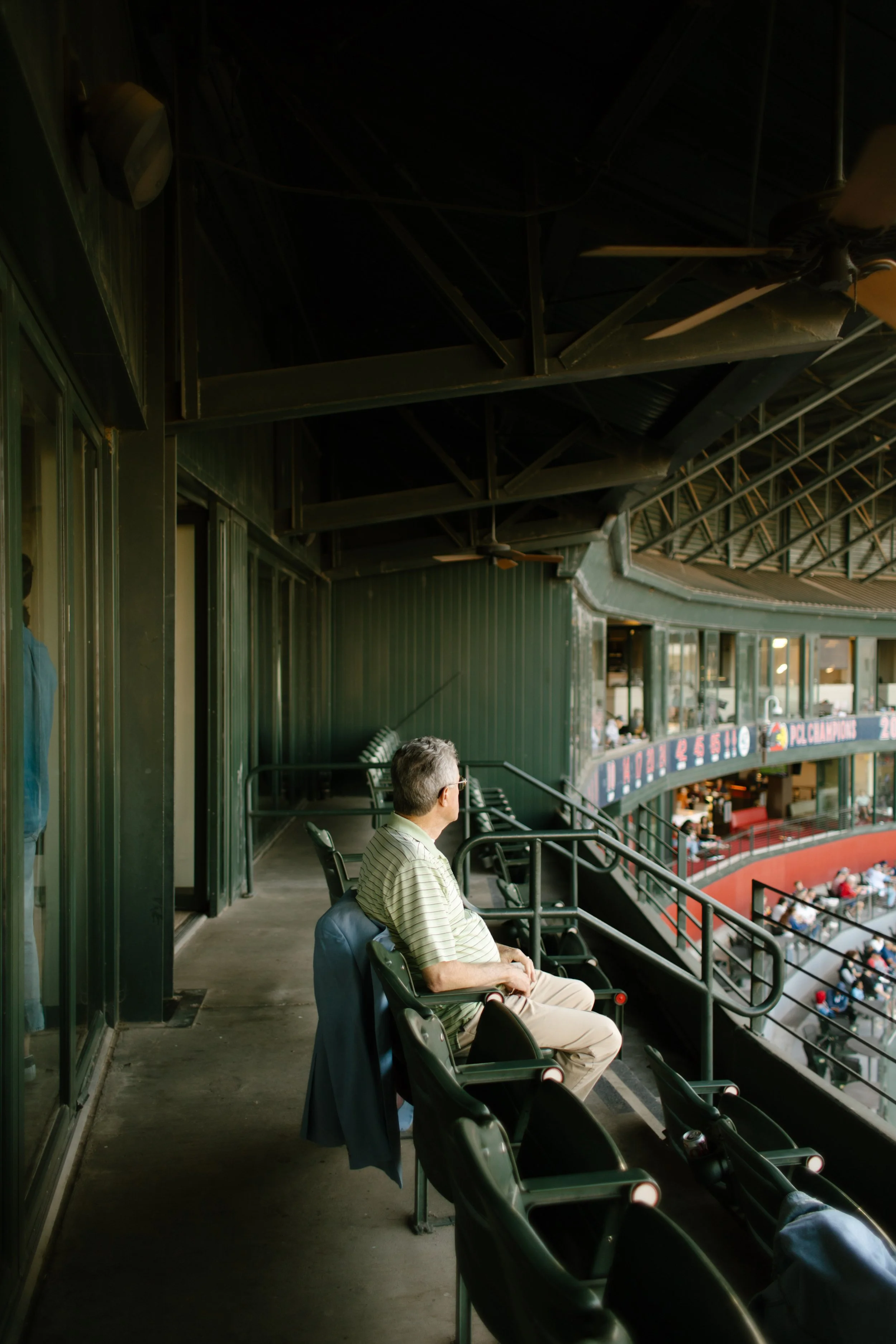 A man sits alone in a quiet section of upper-level stadium seats, looking out toward the baseball field.