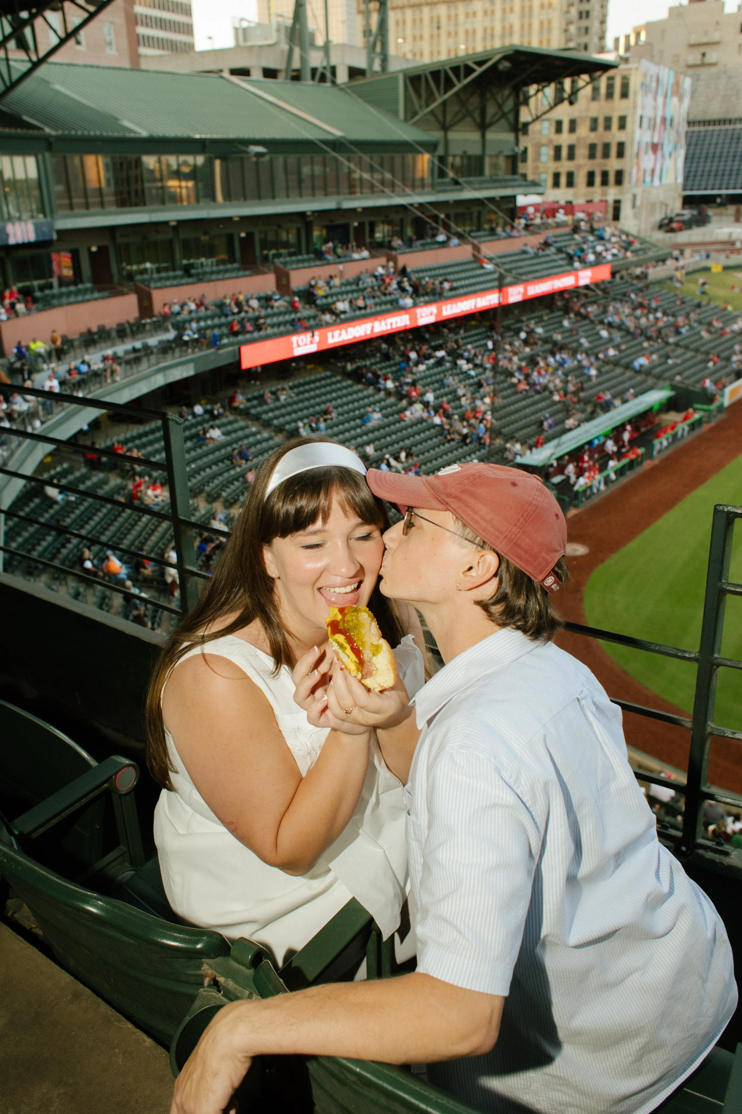 A couple shares a playful kiss on the cheek while enjoying a hot dog in their baseball stadium seats, inspiring fun engagement photos centered on shared experiences.