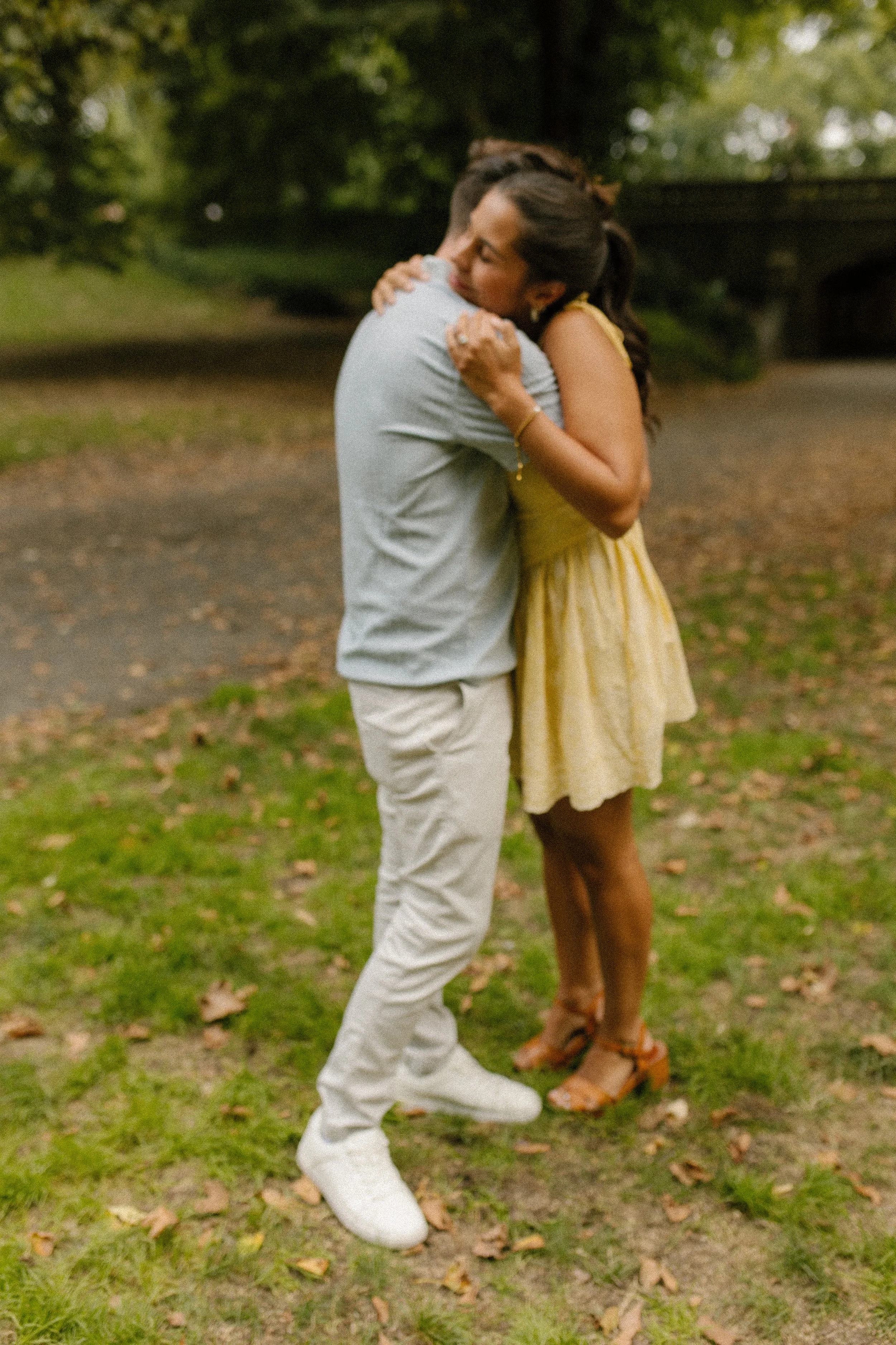 Chris and Sienna embrace on a grassy pathway in Central Park, surrounded by trees shortly after getting engaged.