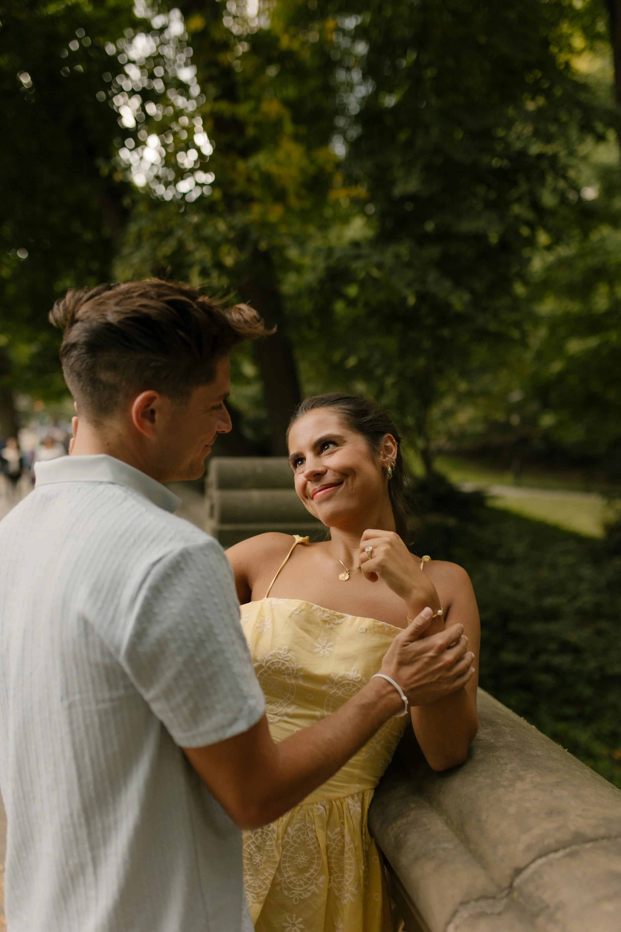 Sienna smiles at Chris while leaning against a stone railing, showing off her engagement ring as they enjoy a quiet moment together.
