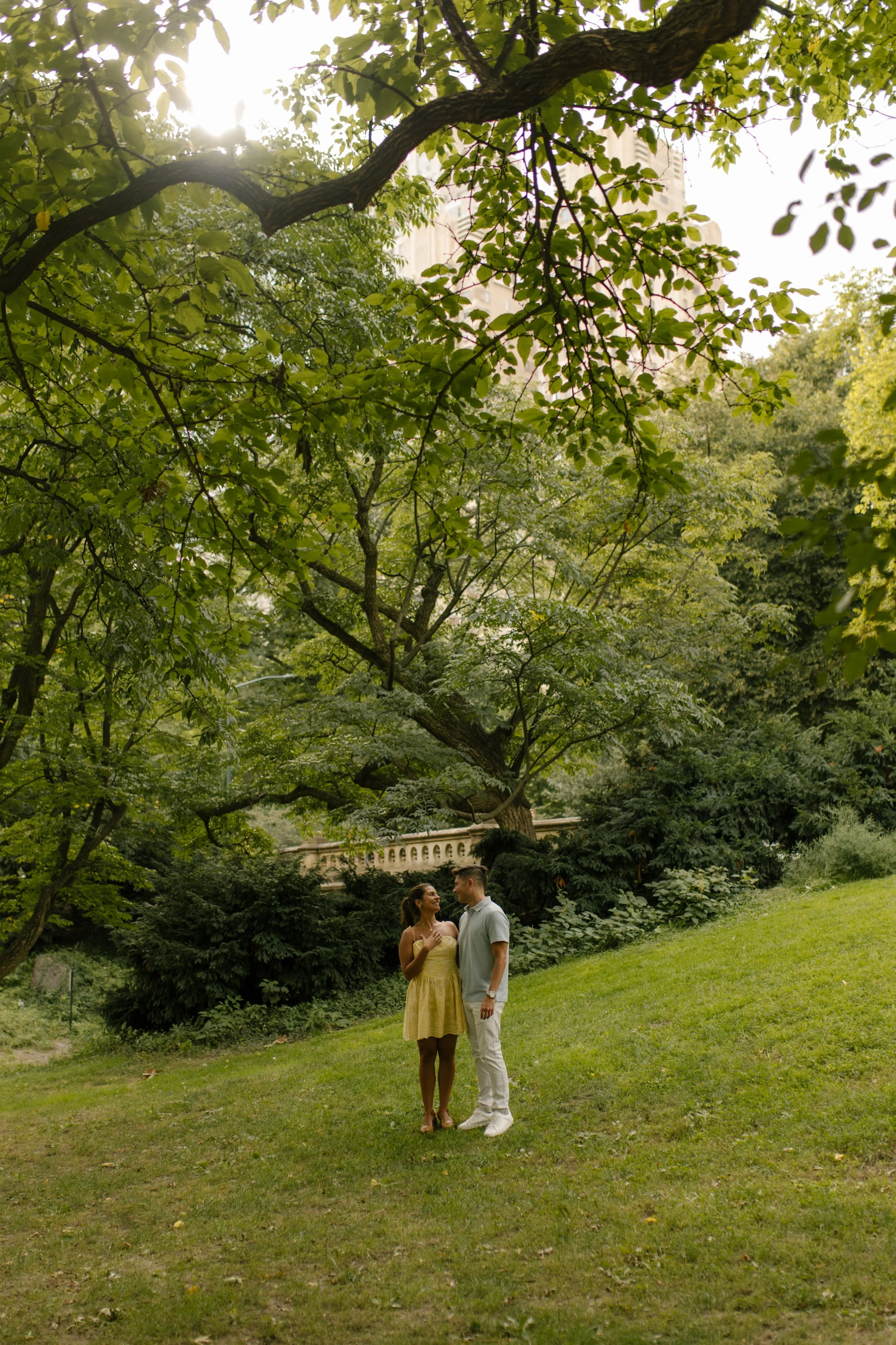 Chris and Sienna stand together on a grassy hillside beneath leafy trees during their intimate new york proposal celebration in Central Park.