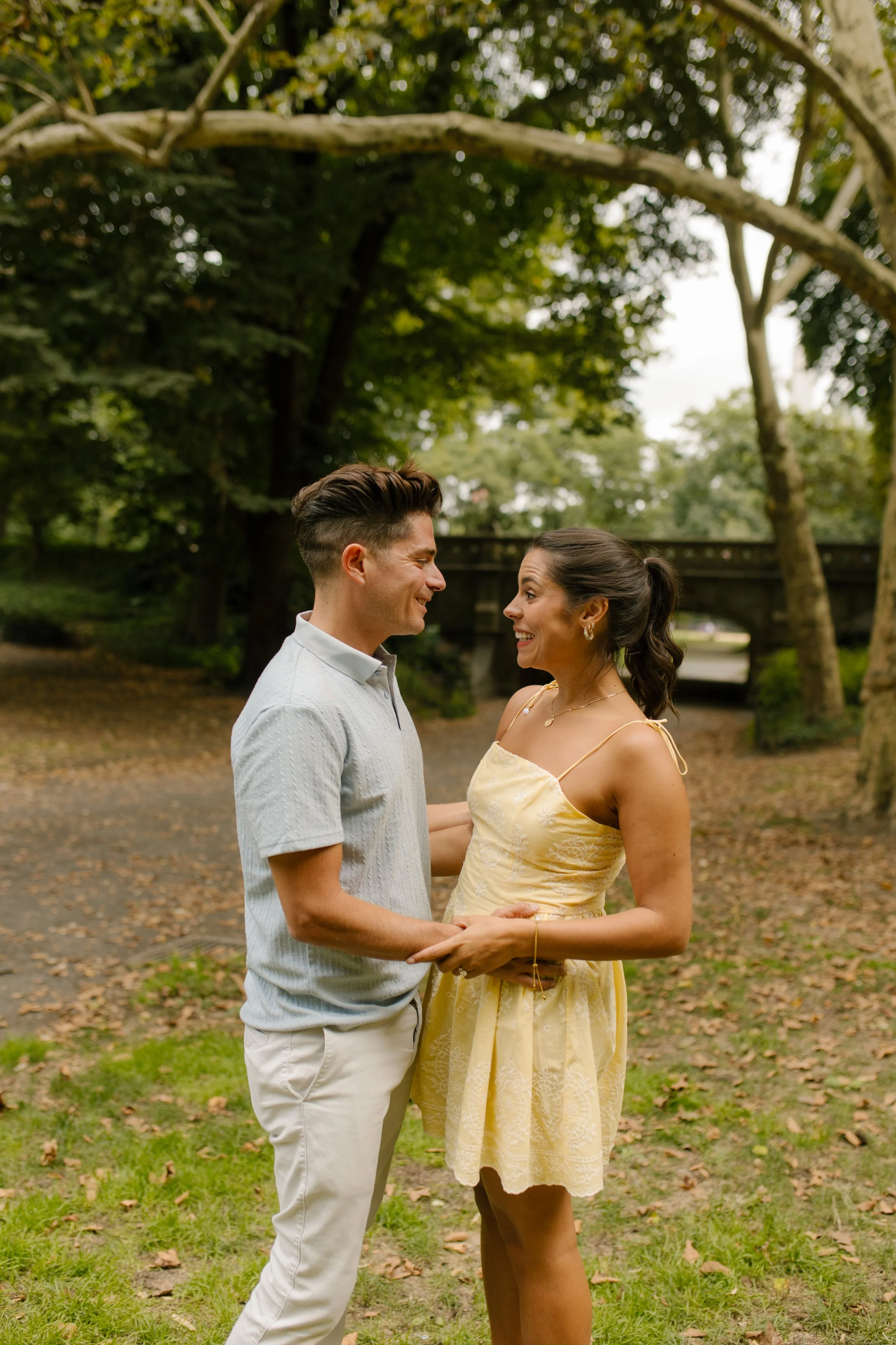 Chris and Sienna hold hands and smile at each other beneath the trees in Central Park, celebrating their surprise new york proposal.