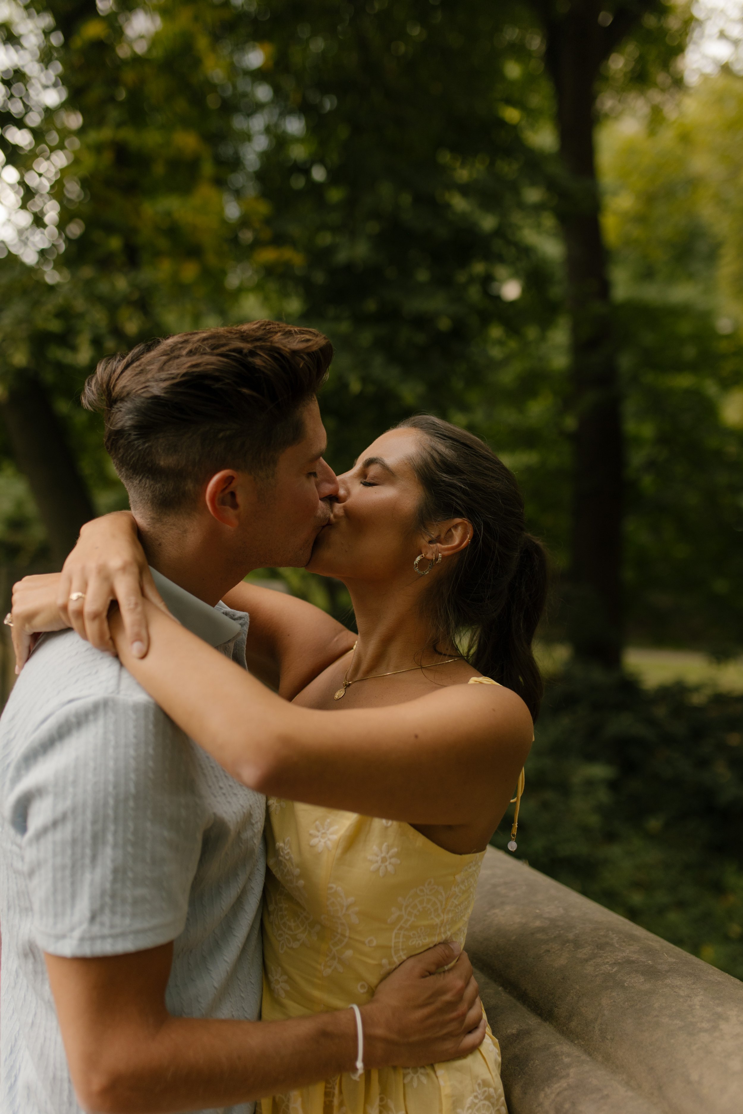 Sienna wraps her arms around Chris's shoulders as they kiss beside a stone railing surrounded by greenery in Central Park.