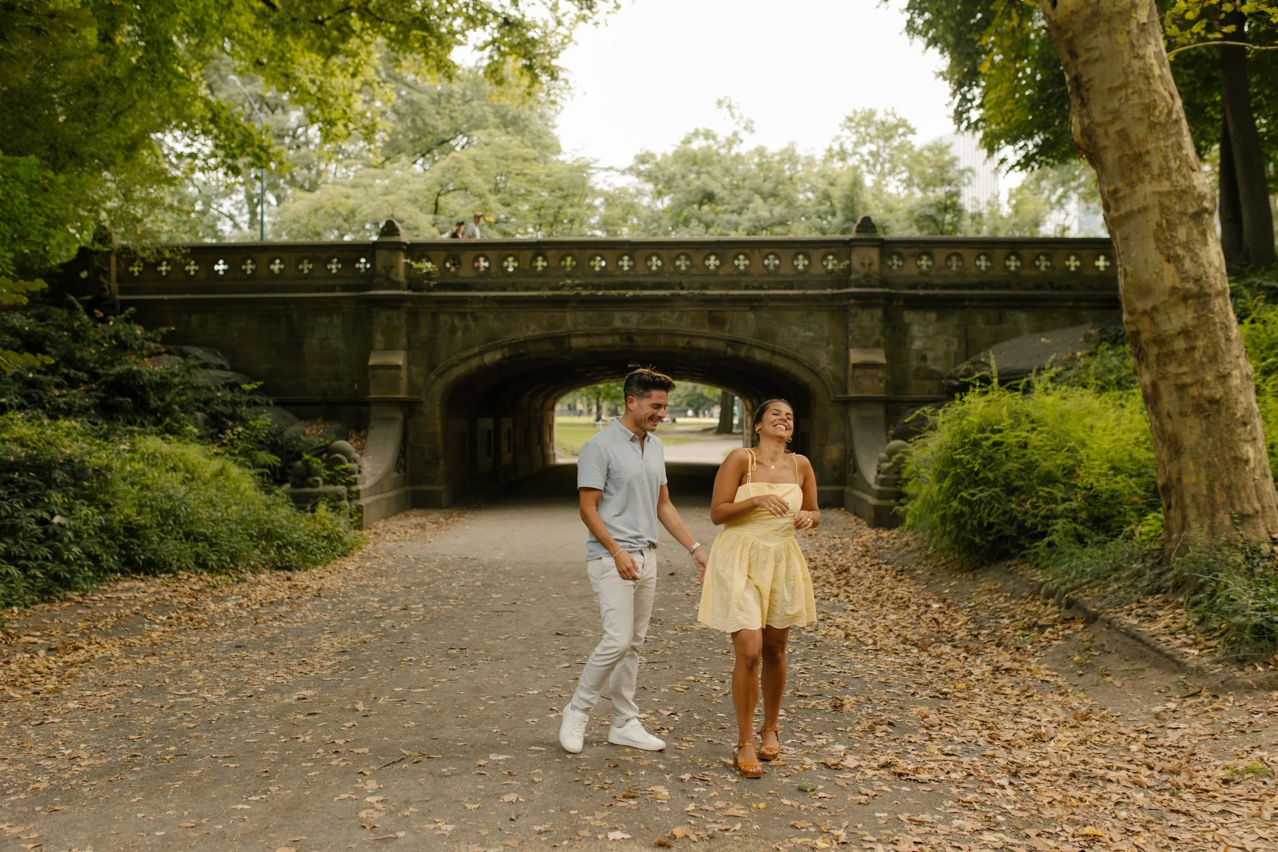 Chris and Sienna walk together along a leaf-covered Central Park pathway, smiling beneath a historic stone bridge after getting engaged.