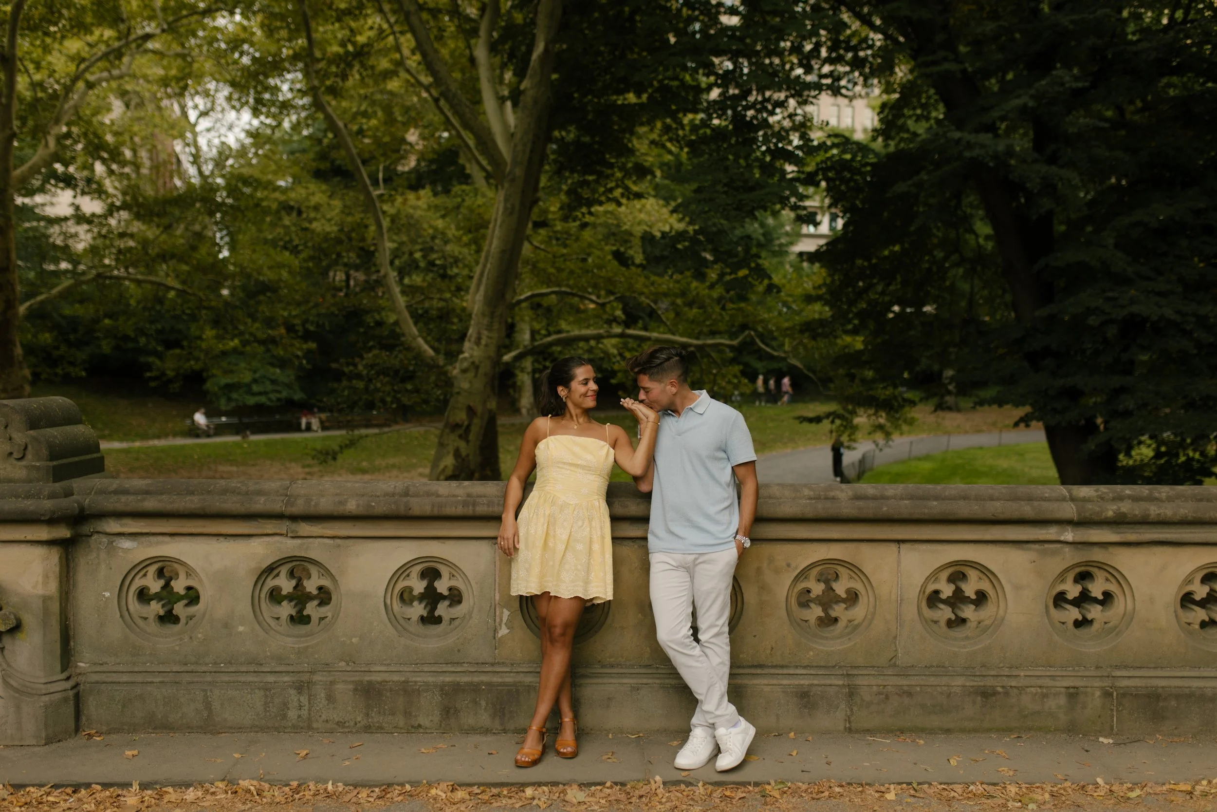 Chris kisses Sienna's hand as they lean against an ornate stone railing during their romantic new york proposal portraits in Central Park.
