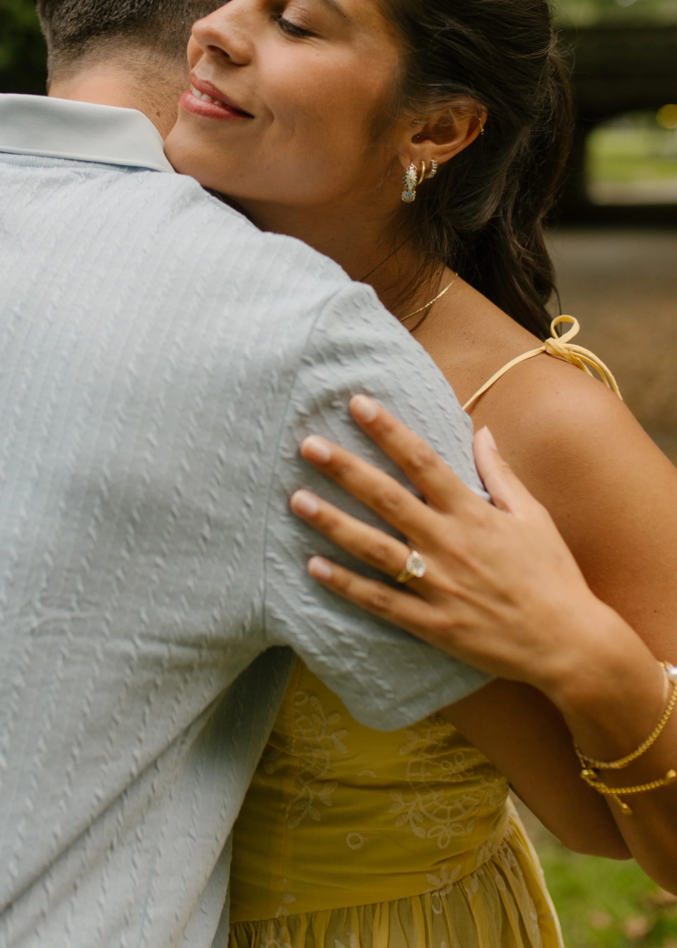 Close-up of Sienna's diamond engagement ring as she embraces Chris, smiling with her eyes closed while wearing a pale yellow dress.