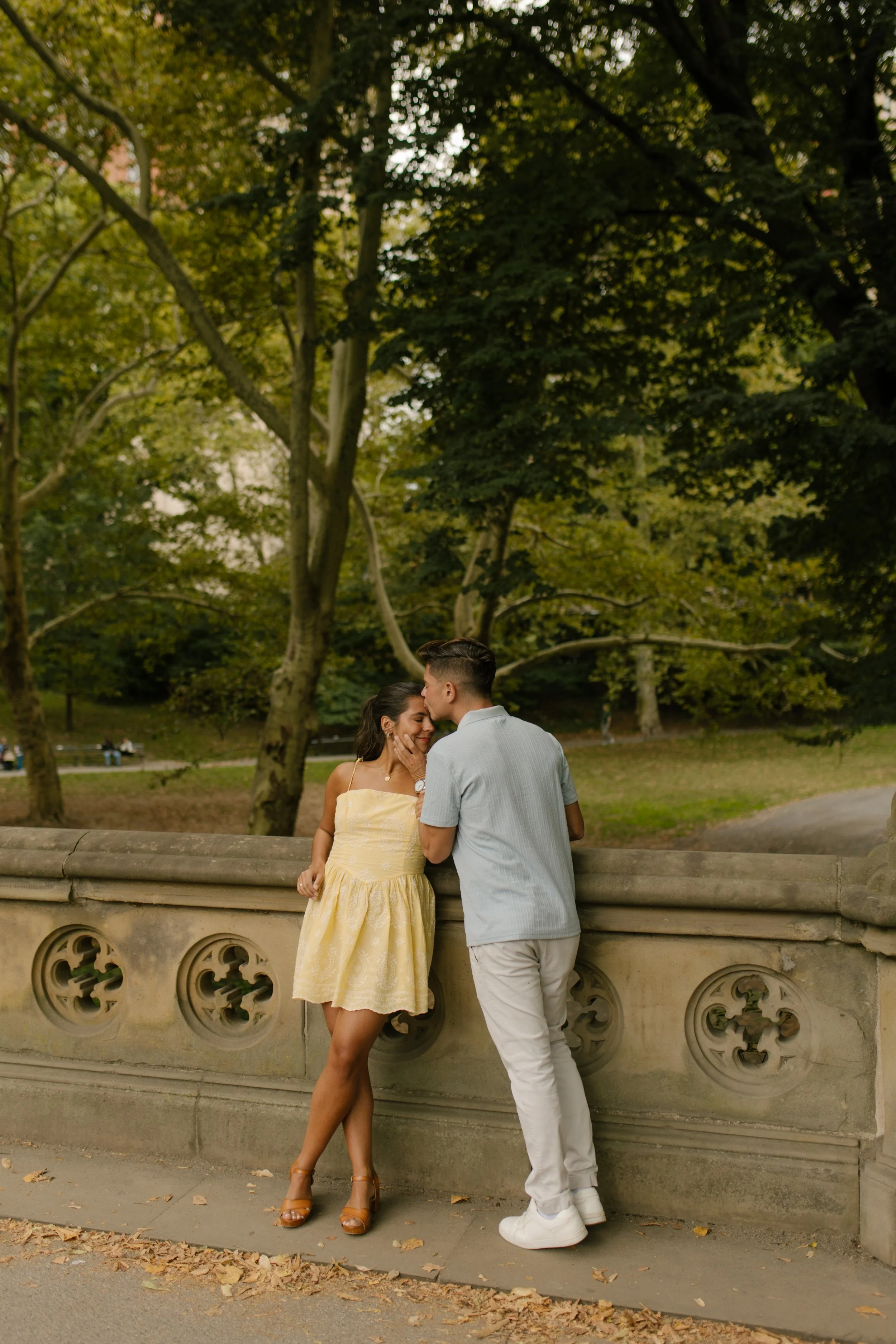 Chris leans in to kiss Sienna as they stand beside an ornate stone railing during their new york proposal portraits in Central Park.