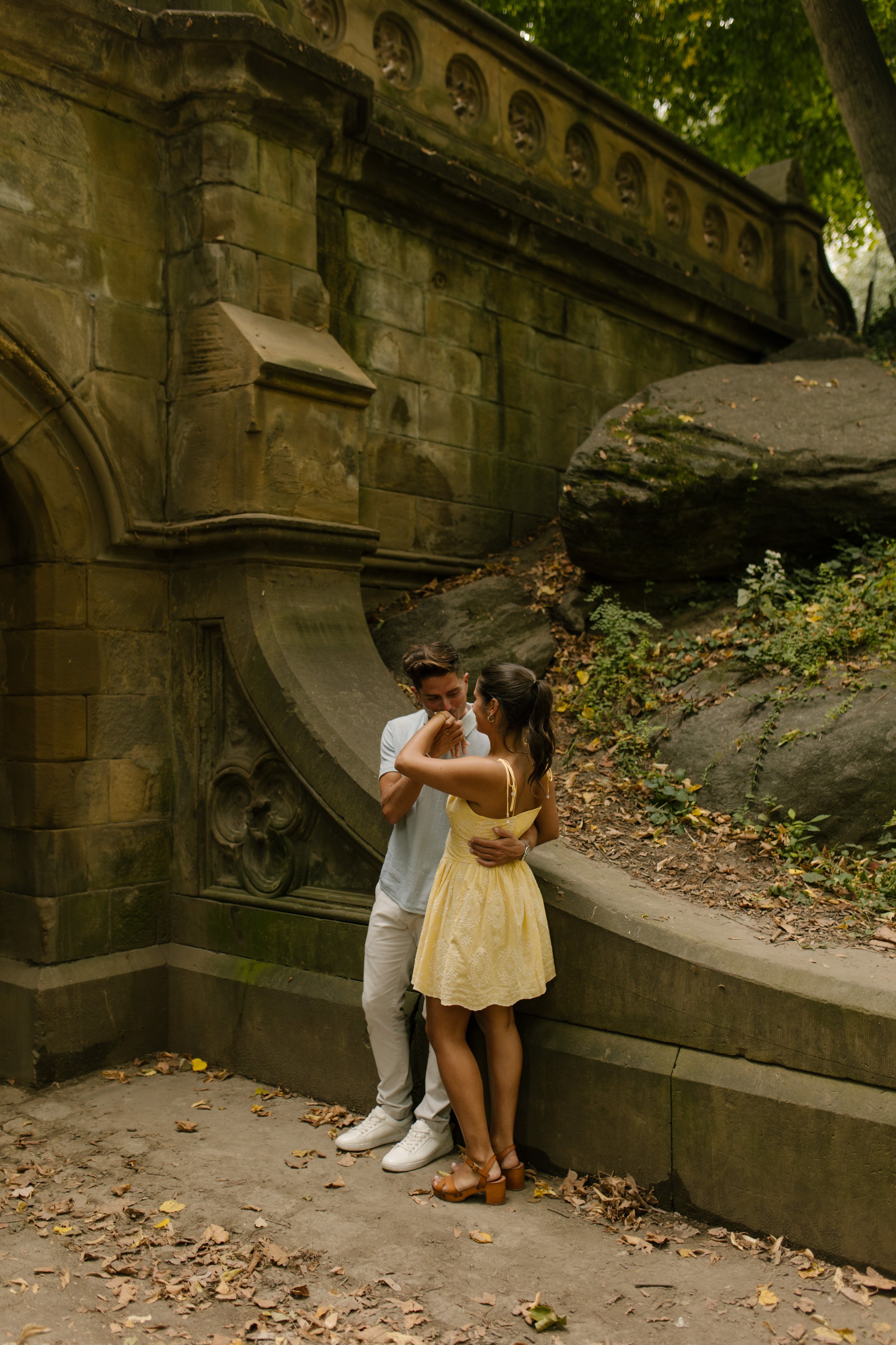 Chris and Sienna embrace beside a curved stone wall beneath a historic Central Park bridge, surrounded by rocks and greenery.