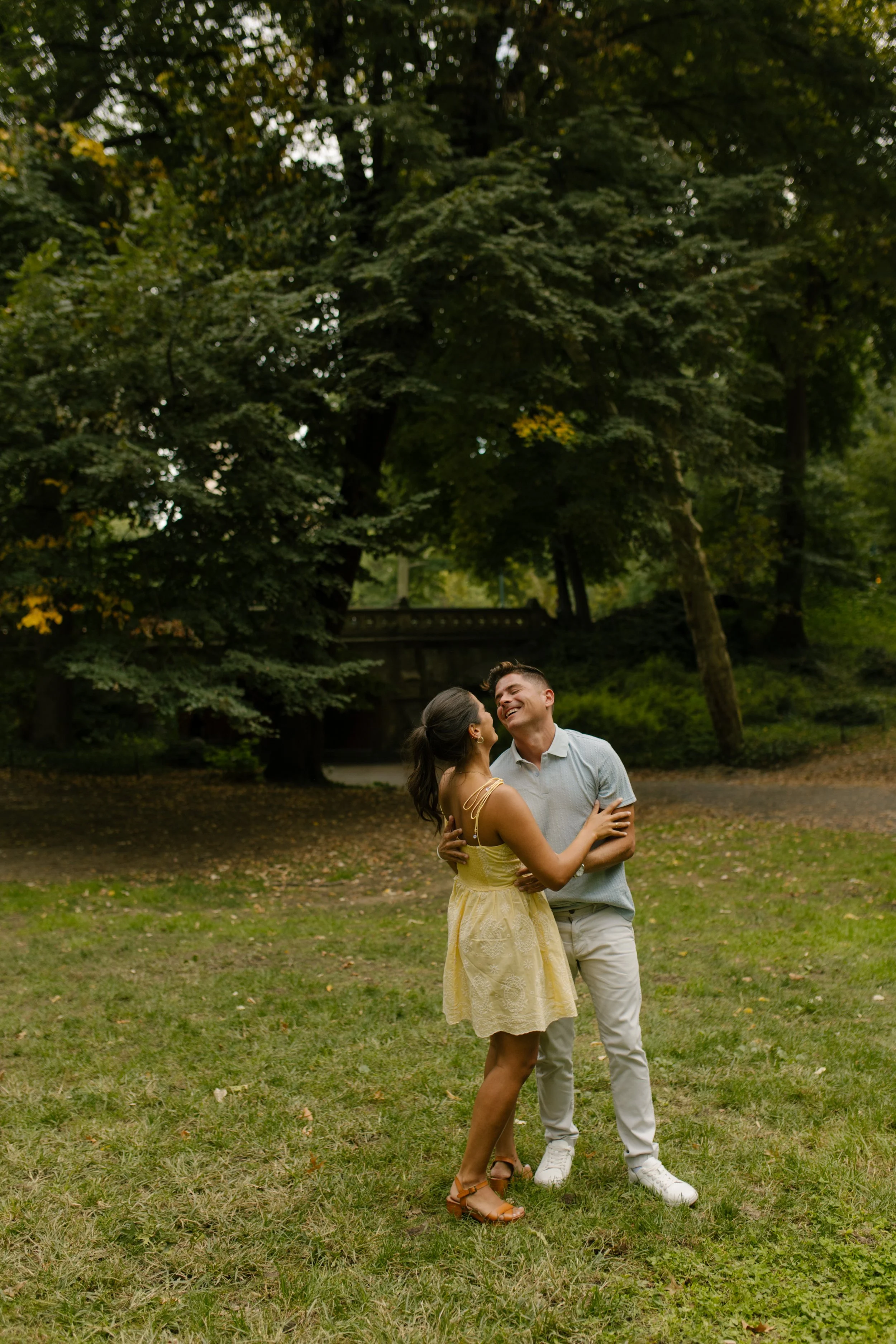 Sienna and Chris laugh together on a grassy lawn in Central Park, with Sienna wearing a short yellow dress and Chris dressed in a light blue polo and white pants.