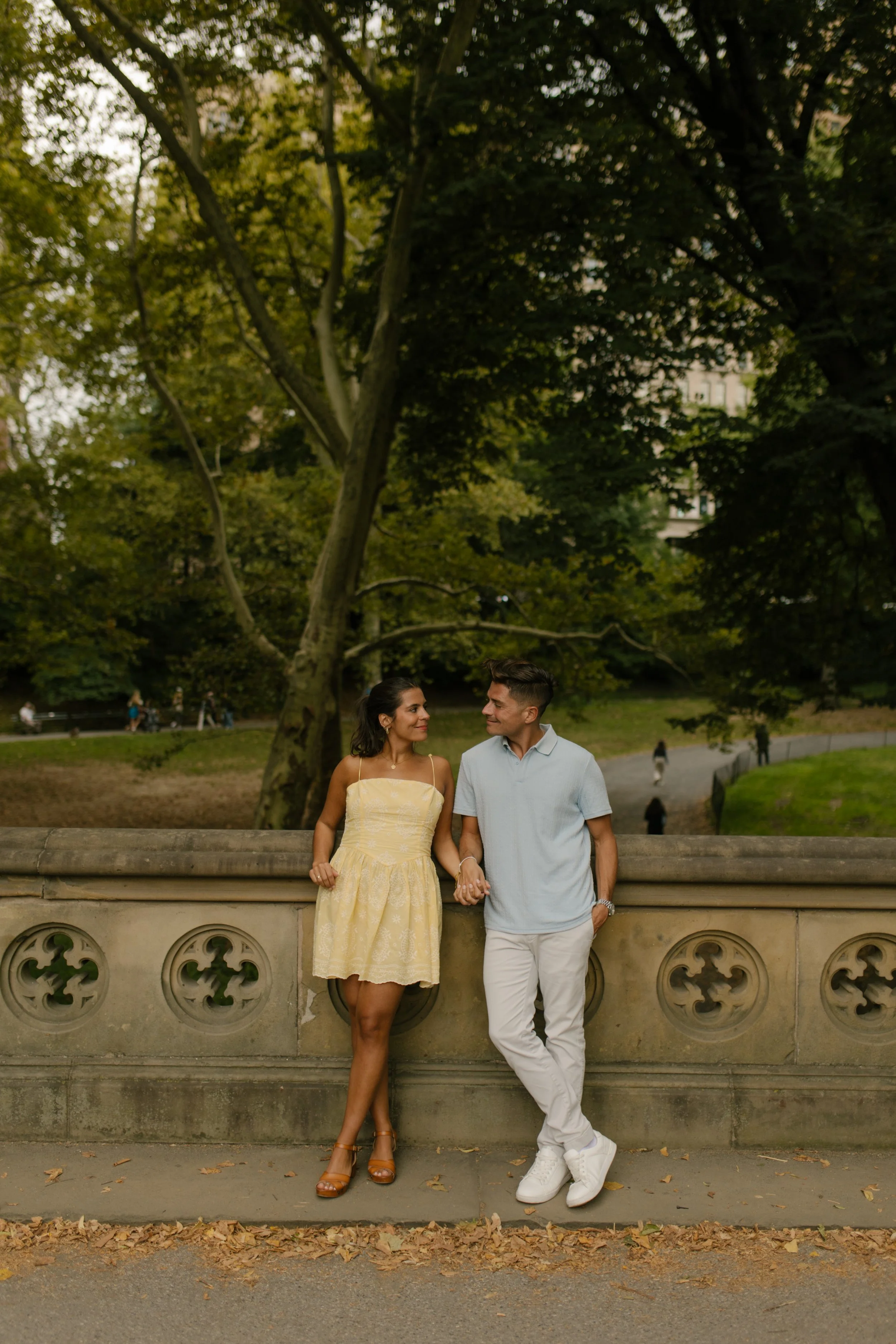 Chris and Sienna hold hands and smile at each other while leaning against a decorative stone railing surrounded by lush Central Park greenery.