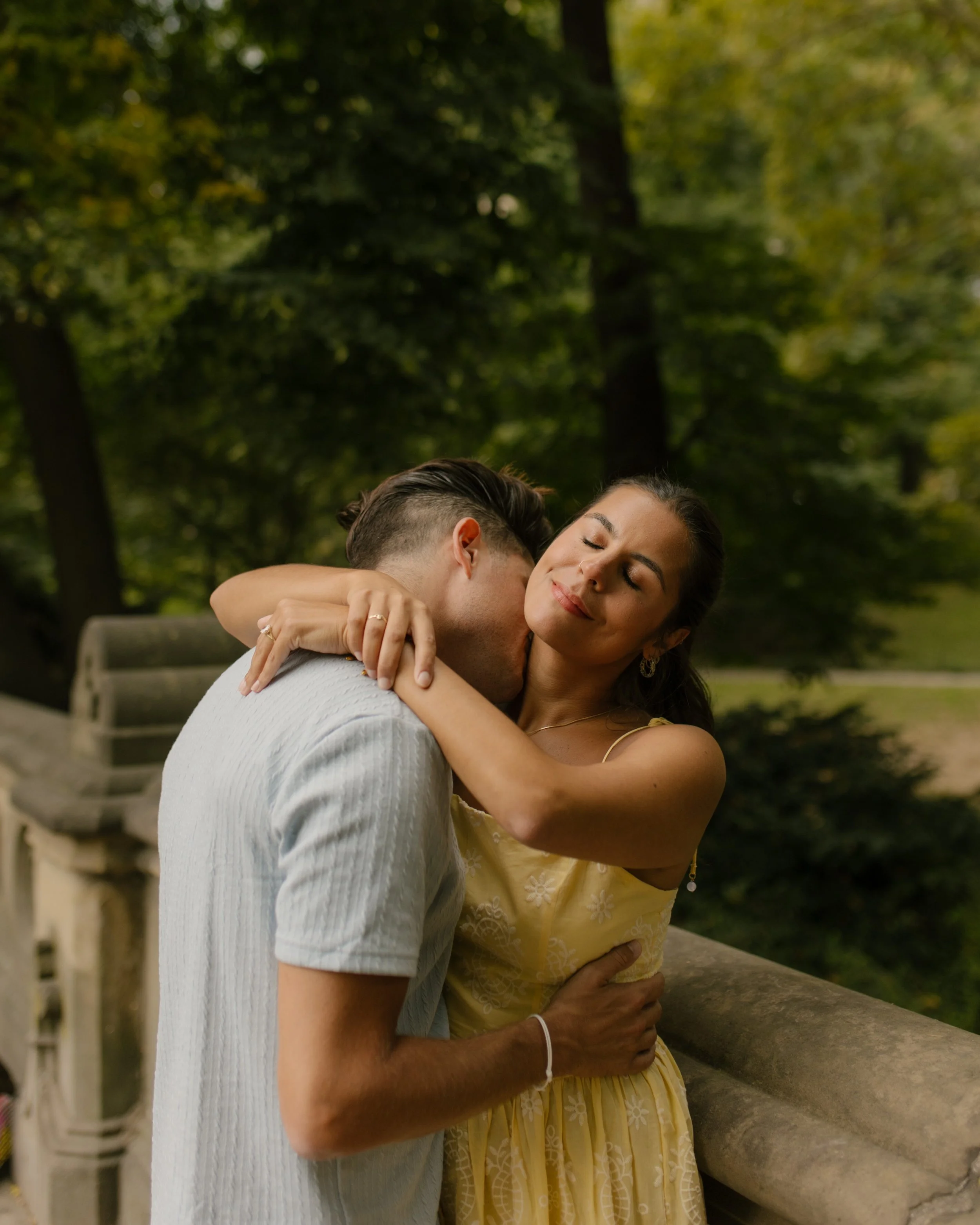 Chris kisses Sienna's neck as she wraps her arms around him beside a stone railing, sharing a romantic moment after their new york proposal in Central Park.