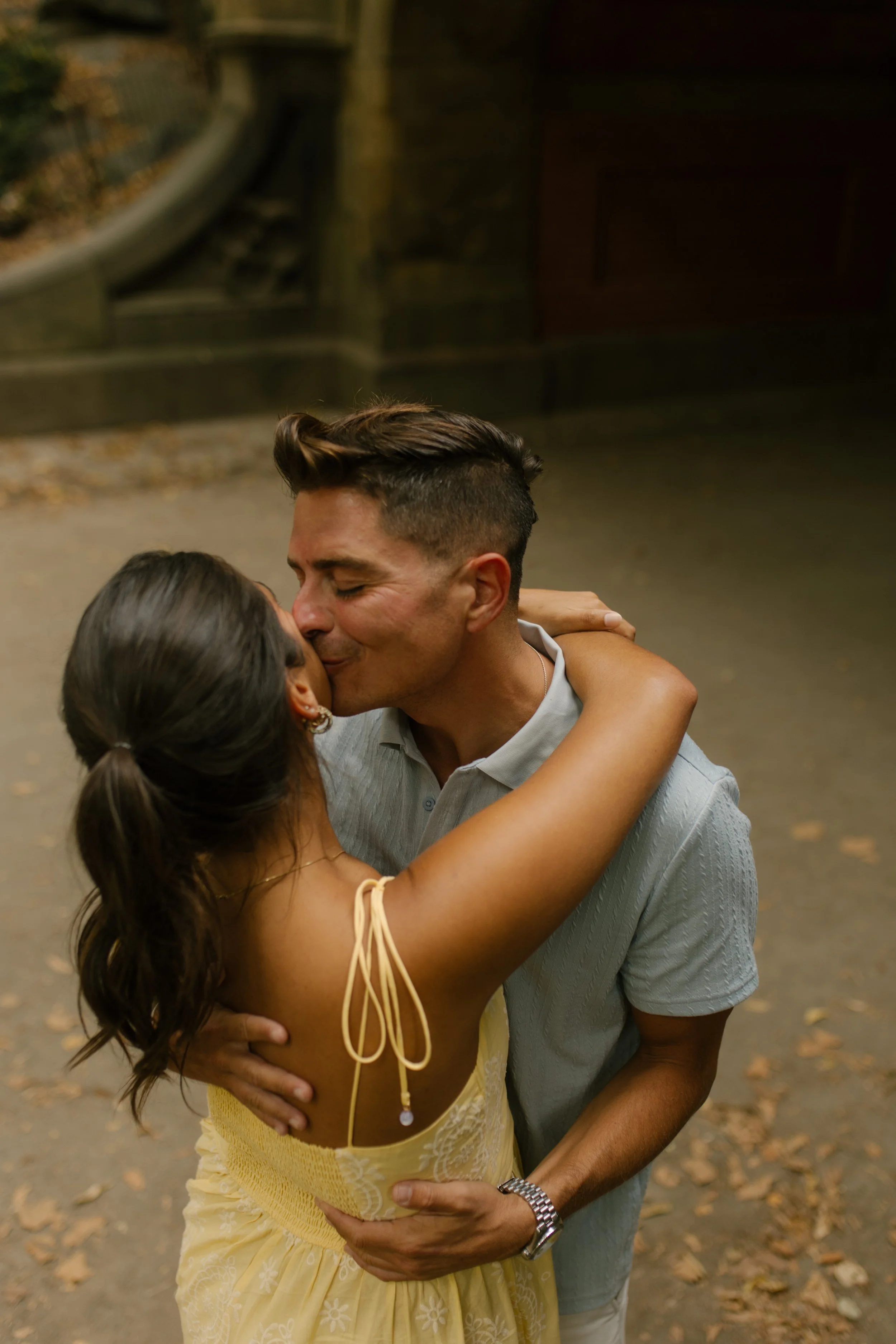 Chris kisses Sienna on the cheek as she wraps her arms around his shoulders, celebrating their new york proposal near a stone bridge in Central Park.